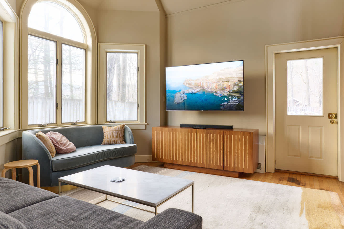 A cozy living area with a wall-mounted flat-screen TV above a wooden media console, and a comfortable gray sofa. An arched bay window with a green velvet sofa is visible to the left, offering a bright seating nook.