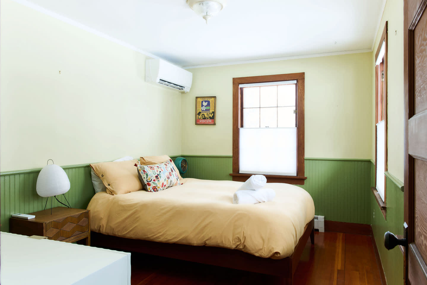 This cozy bedroom features light green walls with wainscoting, a bed with yellow bedding, a wooden nightstand with a white lamp, and a window with light-filtering blinds, showcasing a warm and inviting atmosphere.