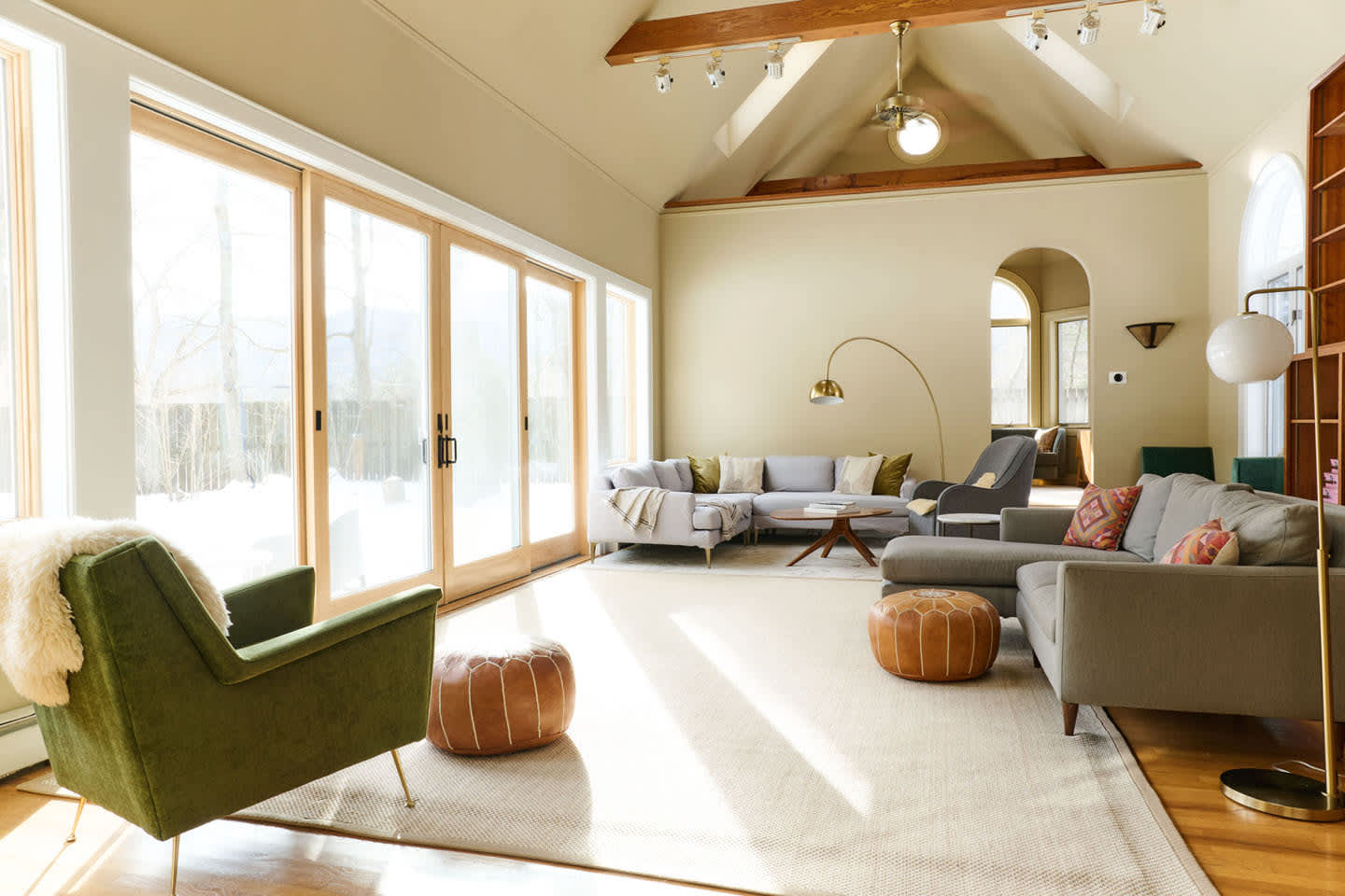 A spacious living room with high ceilings, exposed wooden beams, and a large light-colored sectional sofa. Floor-to-ceiling sliding glass doors offer views of the snowy outdoors, complemented by a mix of modern and mid-century furniture.