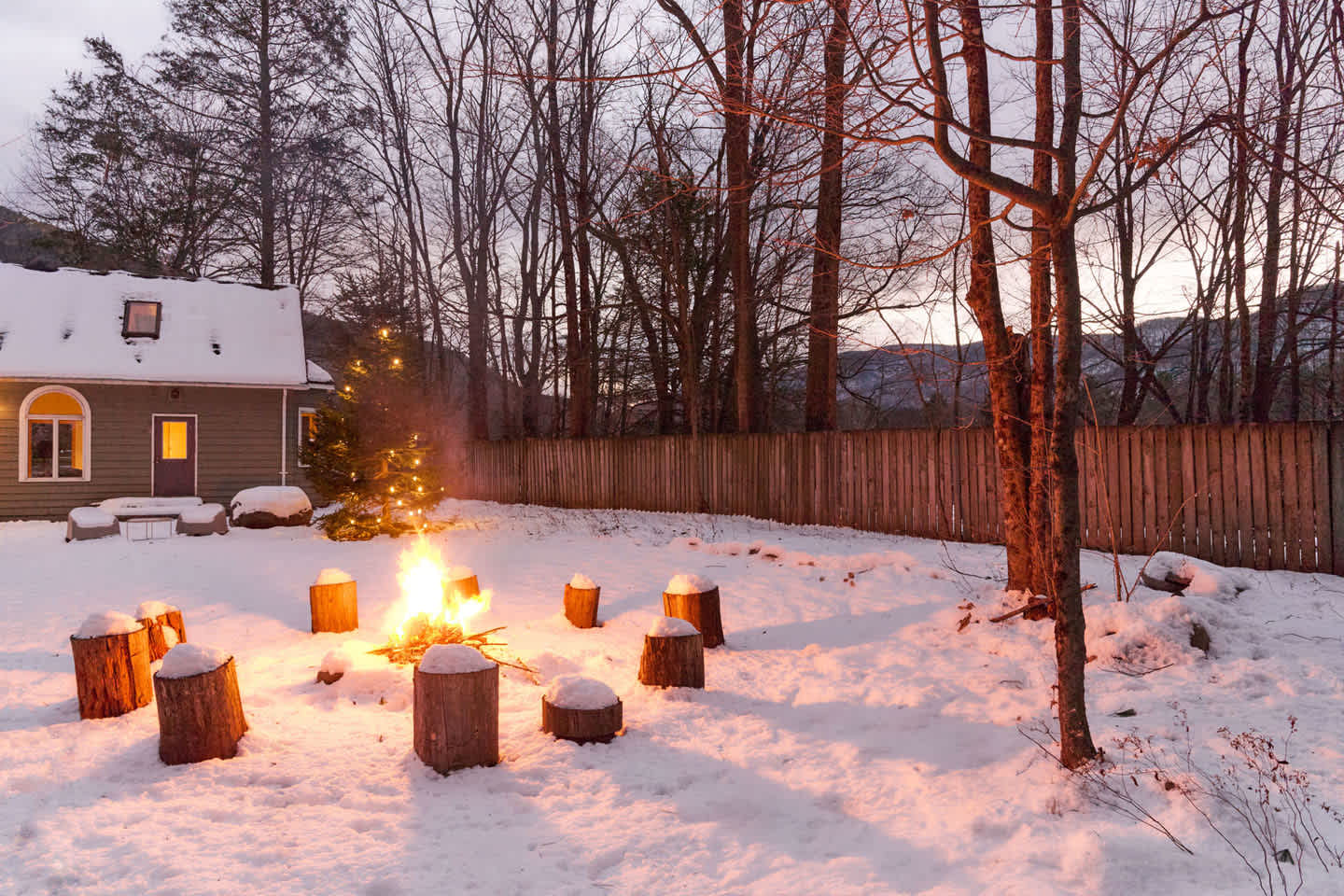 A cozy outdoor fire pit glows brightly in a snow-covered backyard at dusk, surrounded by rustic tree stump seating. A house with a lit Christmas tree is visible in the background, framed by bare winter trees.