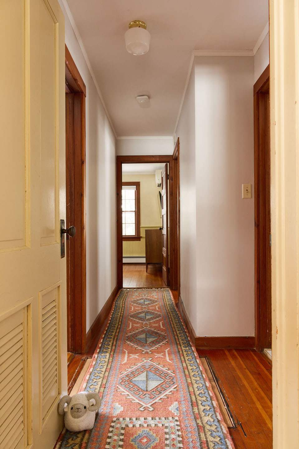 A narrow hallway features light-colored walls and polished hardwood floors, adorned with a colorful patterned runner rug. Wooden door frames and doors are visible, leading to other rooms within the house.