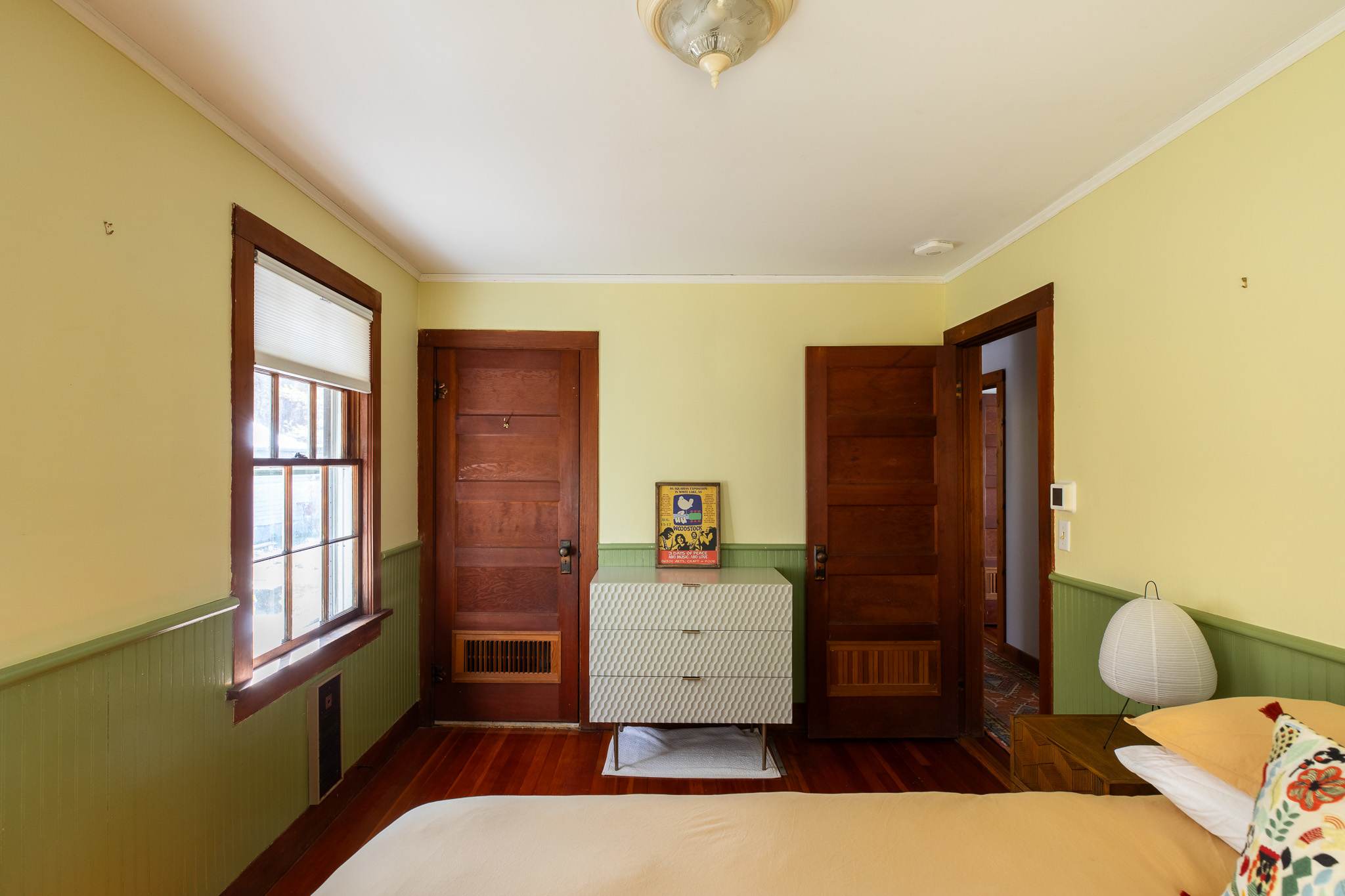 A cozy bedroom featuring light yellow walls with green wainscoting, rich wooden window frames and doors, and a modern dresser with a framed picture on top. A bed with a light-colored duvet is visible in the foreground.
