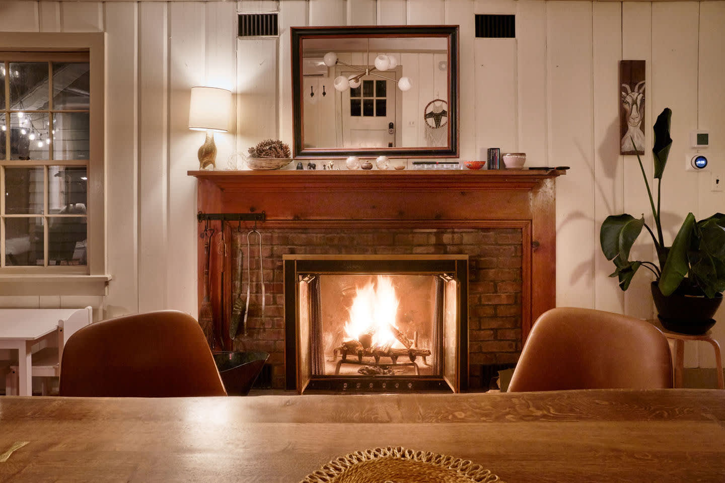 A cozy dining area features a brick fireplace with a wooden mantel, adorned with a large mirror and decorative items. The warm glow of the fire is visible, creating an inviting atmosphere.
