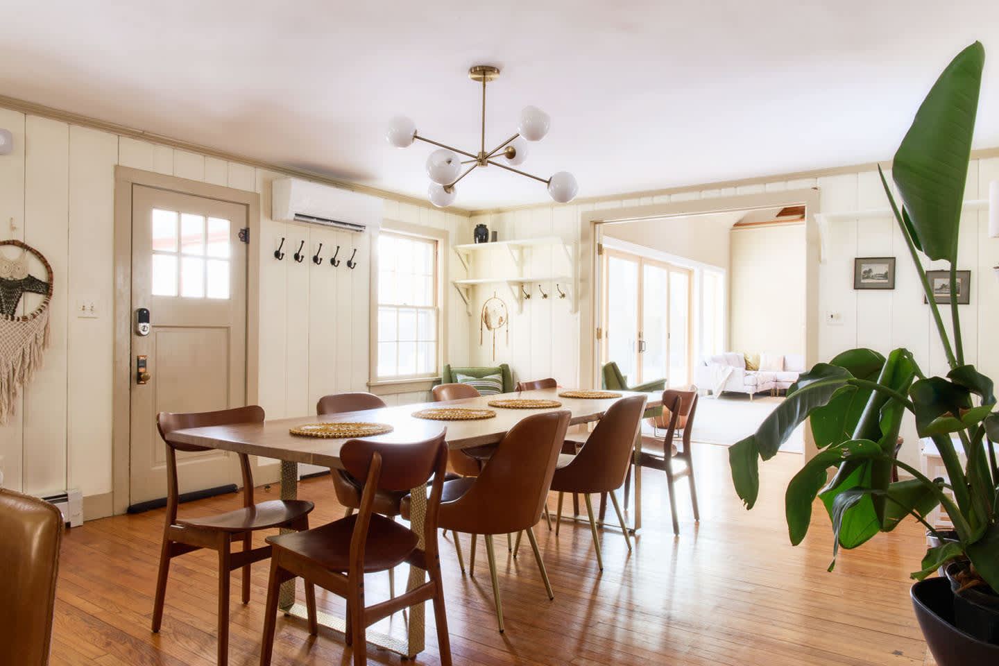 A bright dining area features a long wooden table and chairs, illuminated by a modern Sputnik chandelier and set on hardwood floors. White vertical paneling lines the walls, and a large plant adds a touch of greenery.