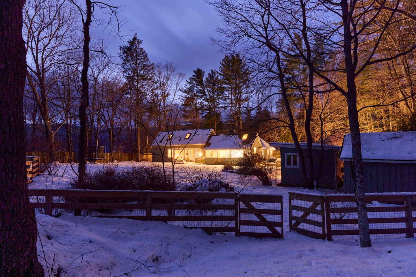 A wide, distant view of the house at dusk, nestled among snow-covered trees, with warm lights emanating from its windows. A rustic wooden fence spans the foreground, and mountains rise in the background.