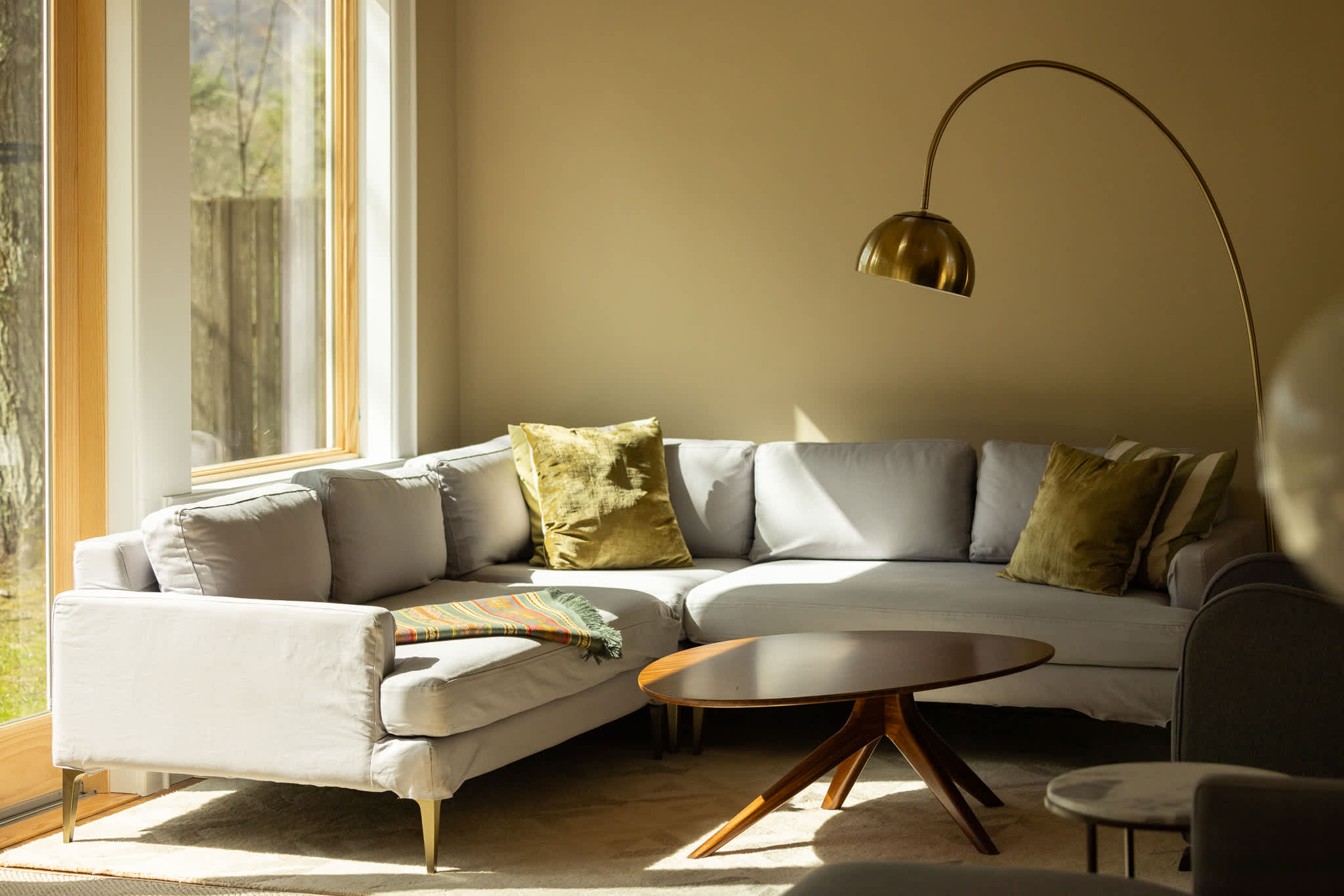 A cozy corner of the living room features a light gray L-shaped sectional sofa with decorative pillows, a modern brass arc floor lamp, and a unique wooden coffee table, all set on a light-colored area rug.