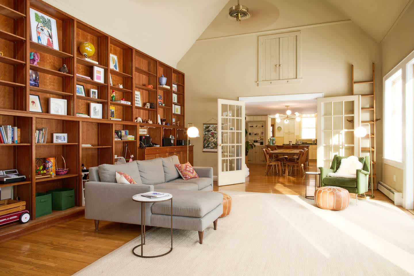 A spacious living room features a floor-to-ceiling wooden bookshelf wall filled with books and decor, a gray sectional sofa with a chaise lounge, and an open view through French doors to a dining area.