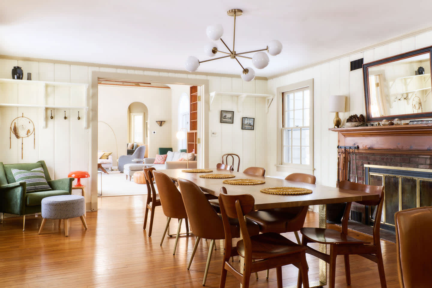 A bright dining room features a long wooden table with seating for eight, illuminated by a modern sputnik chandelier. A traditional fireplace with a dark wood mantel anchors one wall, while hardwood floors extend throughout.