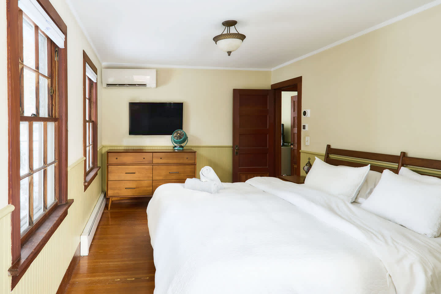 A cozy bedroom with light yellow walls and green wainscoting, featuring a wooden bed frame with white bedding and a wall-mounted TV. Two windows with dark wood trim and an air conditioning unit are visible.