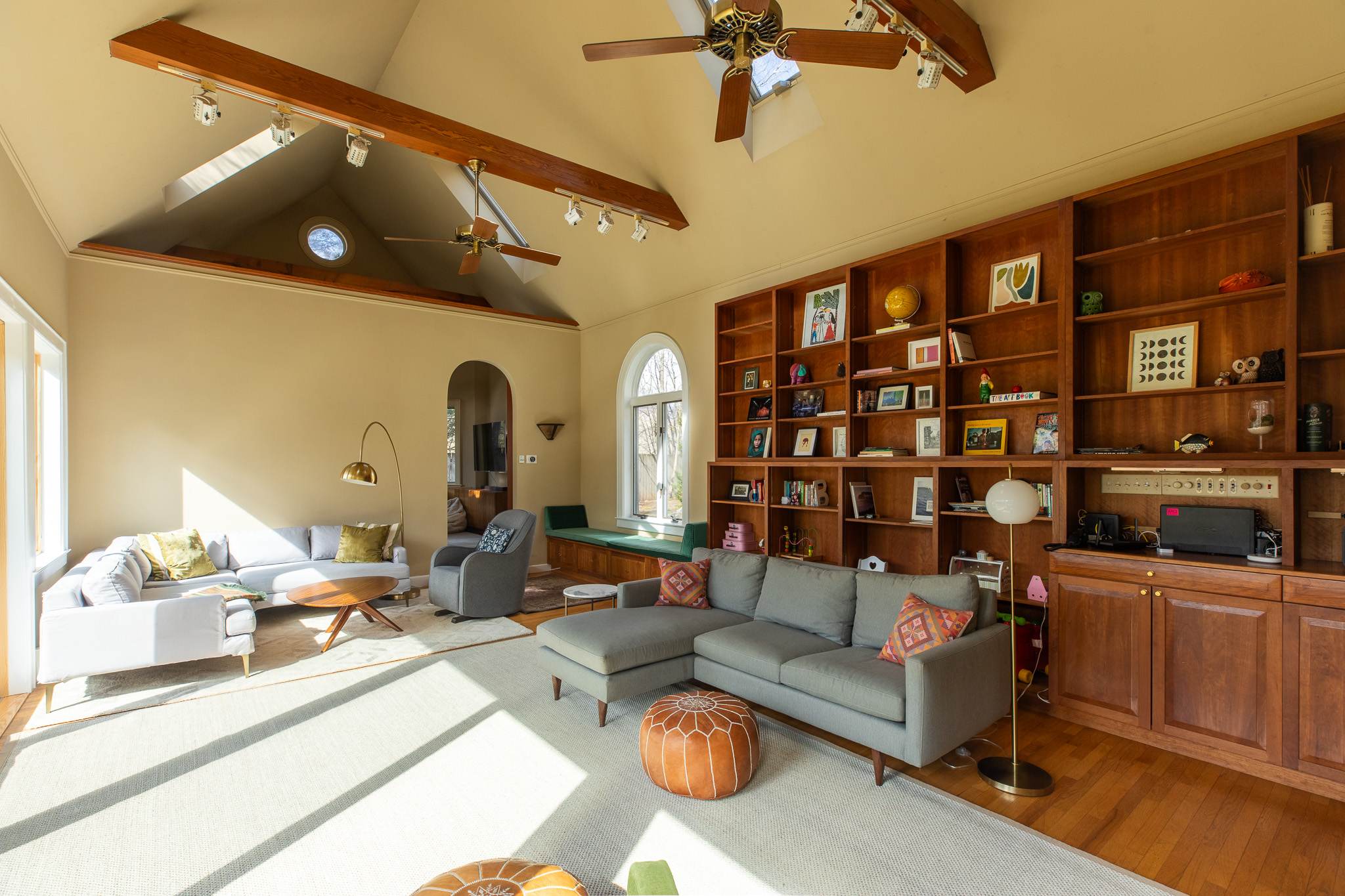 A bright living room with vaulted ceilings and exposed wooden beams features a large wooden built-in bookshelf, a gray sectional sofa, and a light gray sofa, all arranged on a large area rug.
