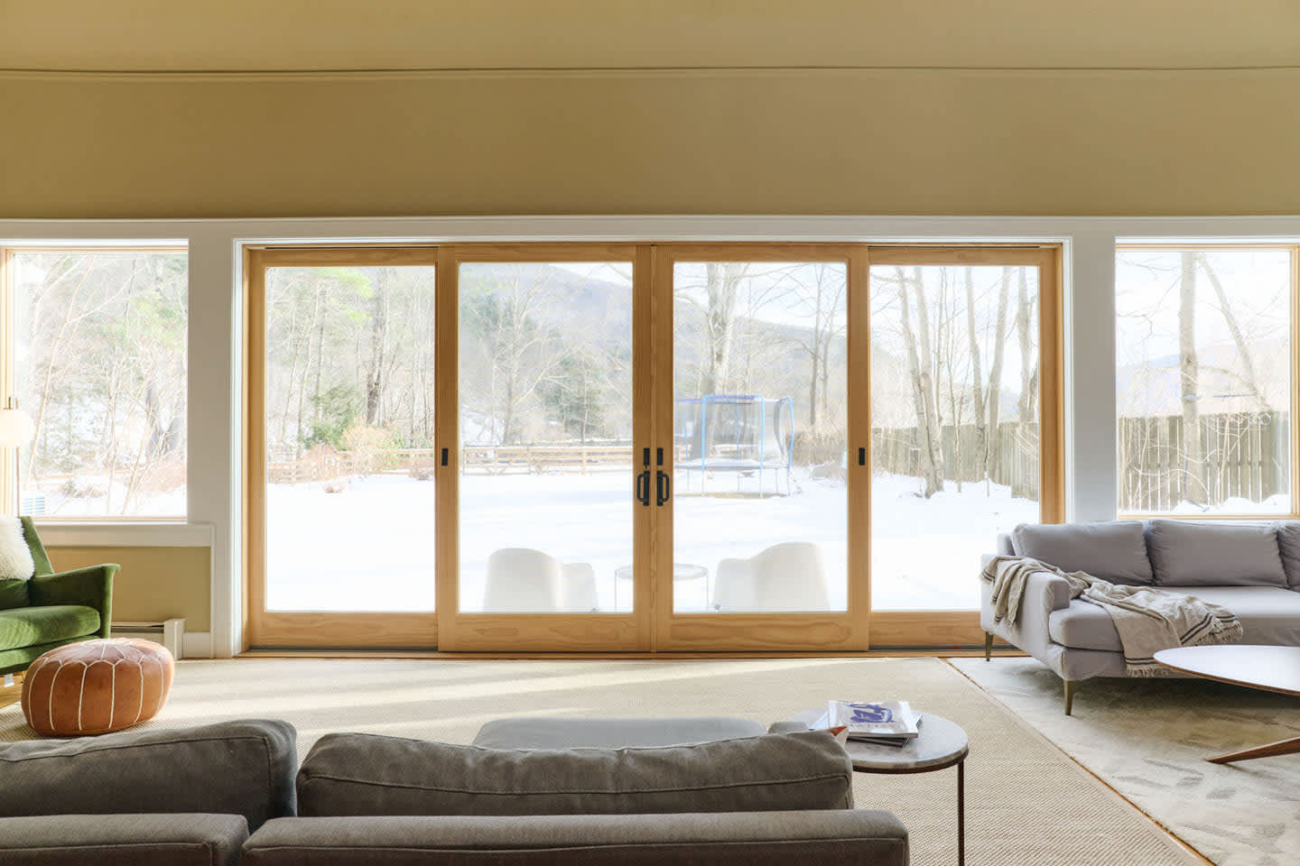 Expansive living room featuring a large light-colored area rug and comfortable sofas, with floor-to-ceiling wooden sliding glass doors opening to a snowy outdoor landscape. Additional windows flank the doors, providing ample natural light.