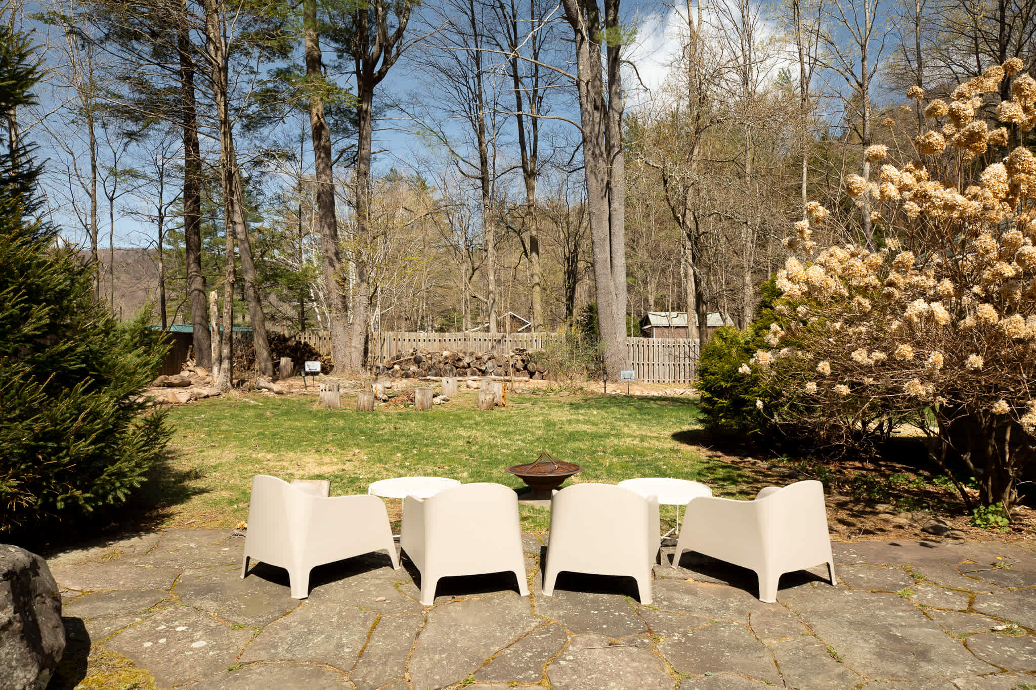 Outdoor patio area with four modern white lounge chairs arranged around a metal fire pit, set on flagstone pavers. The area is surrounded by tall trees and a wooden fence in the background.