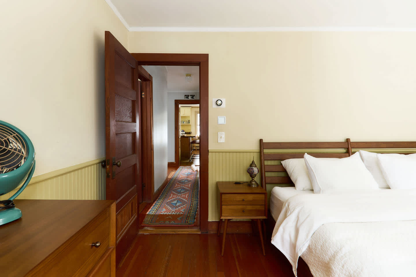A warm bedroom with light yellow walls and green wainscoting, featuring a wooden bed frame and a vintage-style fan on a dresser. An open dark wooden door leads to a hallway with a colorful runner rug.