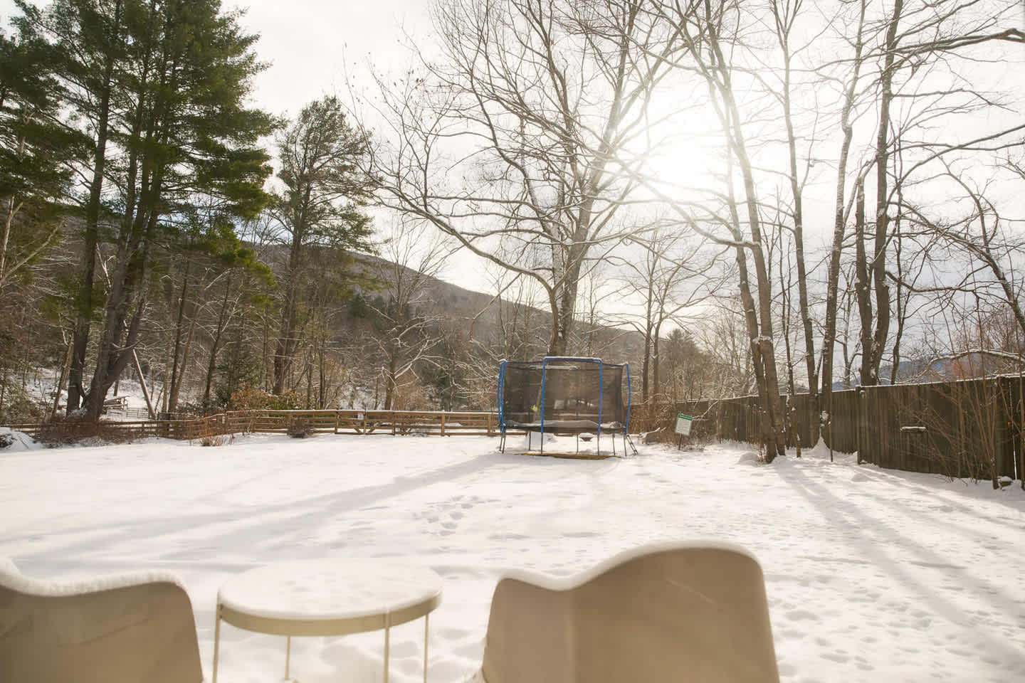 A sunny, snow-covered backyard features a trampoline in the center, surrounded by bare trees and a wooden fence. Distant mountains are visible under a bright, clear sky.