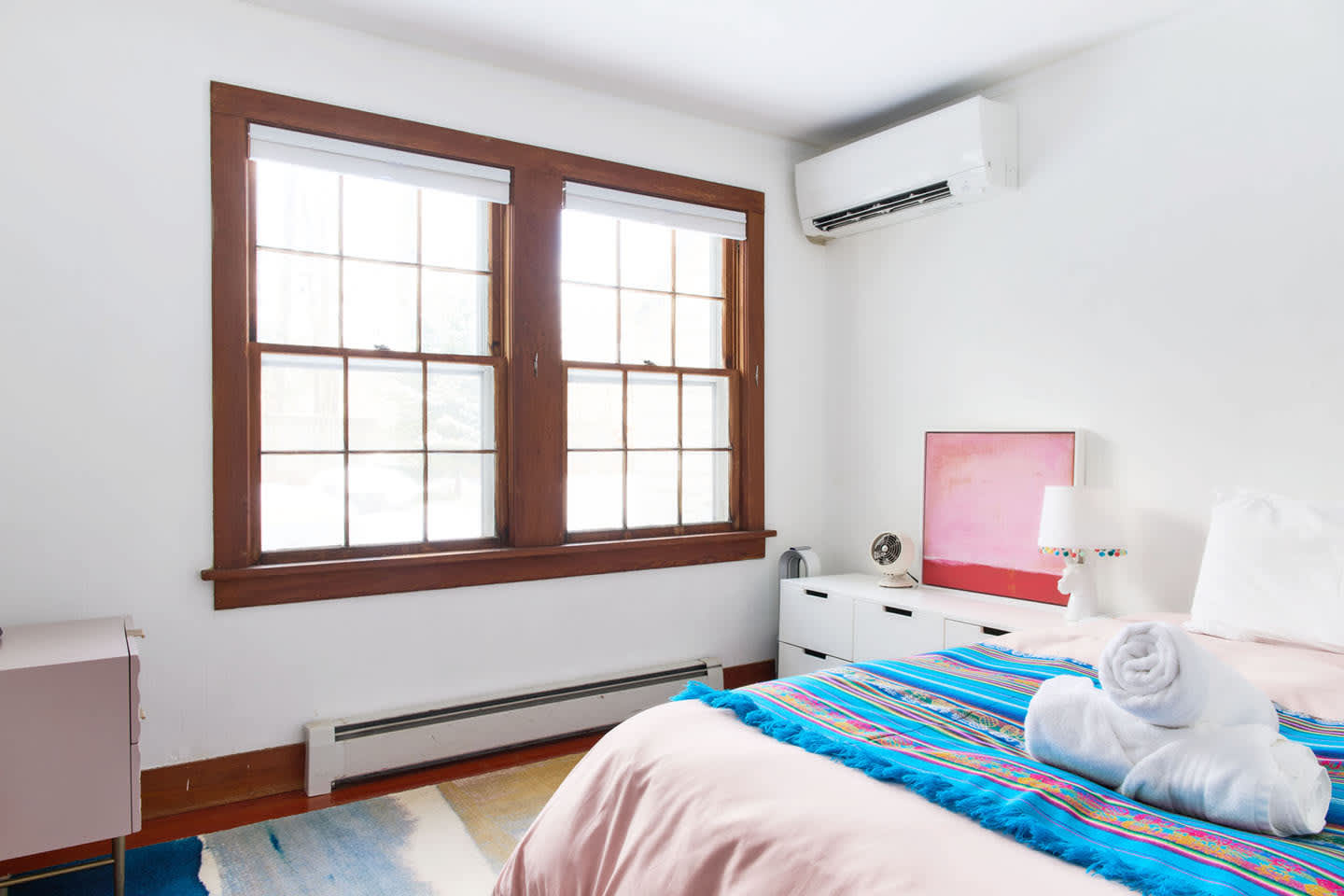 A bright bedroom with white walls, two large windows with dark wood trim, and a white dresser with a pink abstract painting. A bed with a pink duvet and colorful throw blanket is visible, along with an air conditioning unit above the windows.