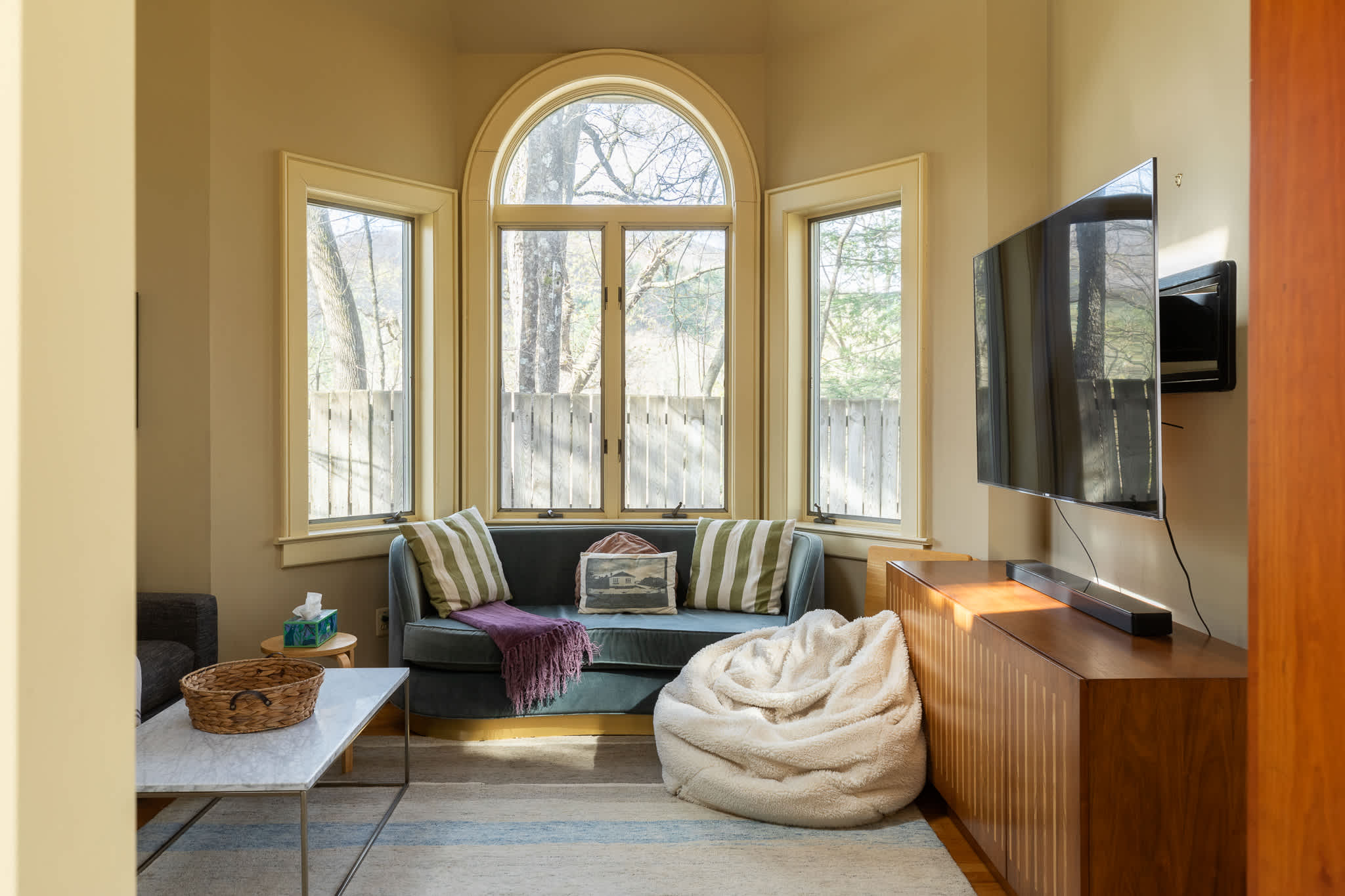 A small, intimate living space features a curved teal velvet sofa with striped pillows, a wall-mounted flat-screen TV, and a wooden media console, all illuminated by natural light from arched and rectangular windows.