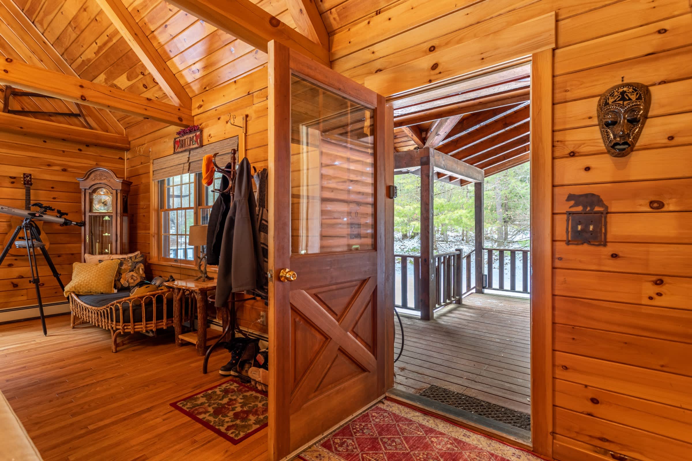 An inviting entryway with natural wood walls and vaulted ceilings features a wooden door opening to an outdoor deck. A coat rack, grandfather clock, and telescope are visible in the background.