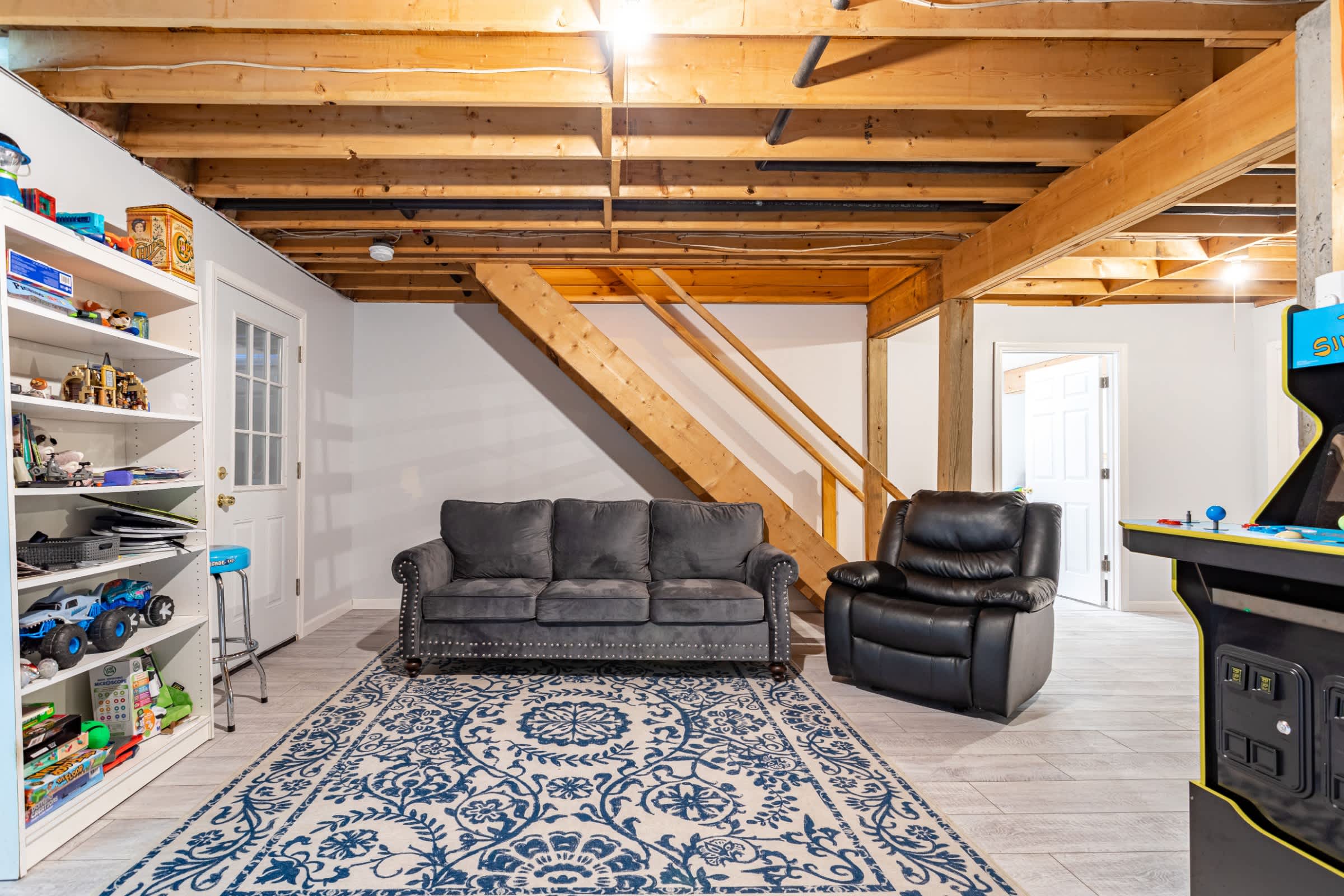 A spacious recreation area with exposed wooden ceiling joists and ductwork, featuring a grey sofa, a black recliner, a blue and white patterned rug, a bookshelf with toys, and an arcade machine.