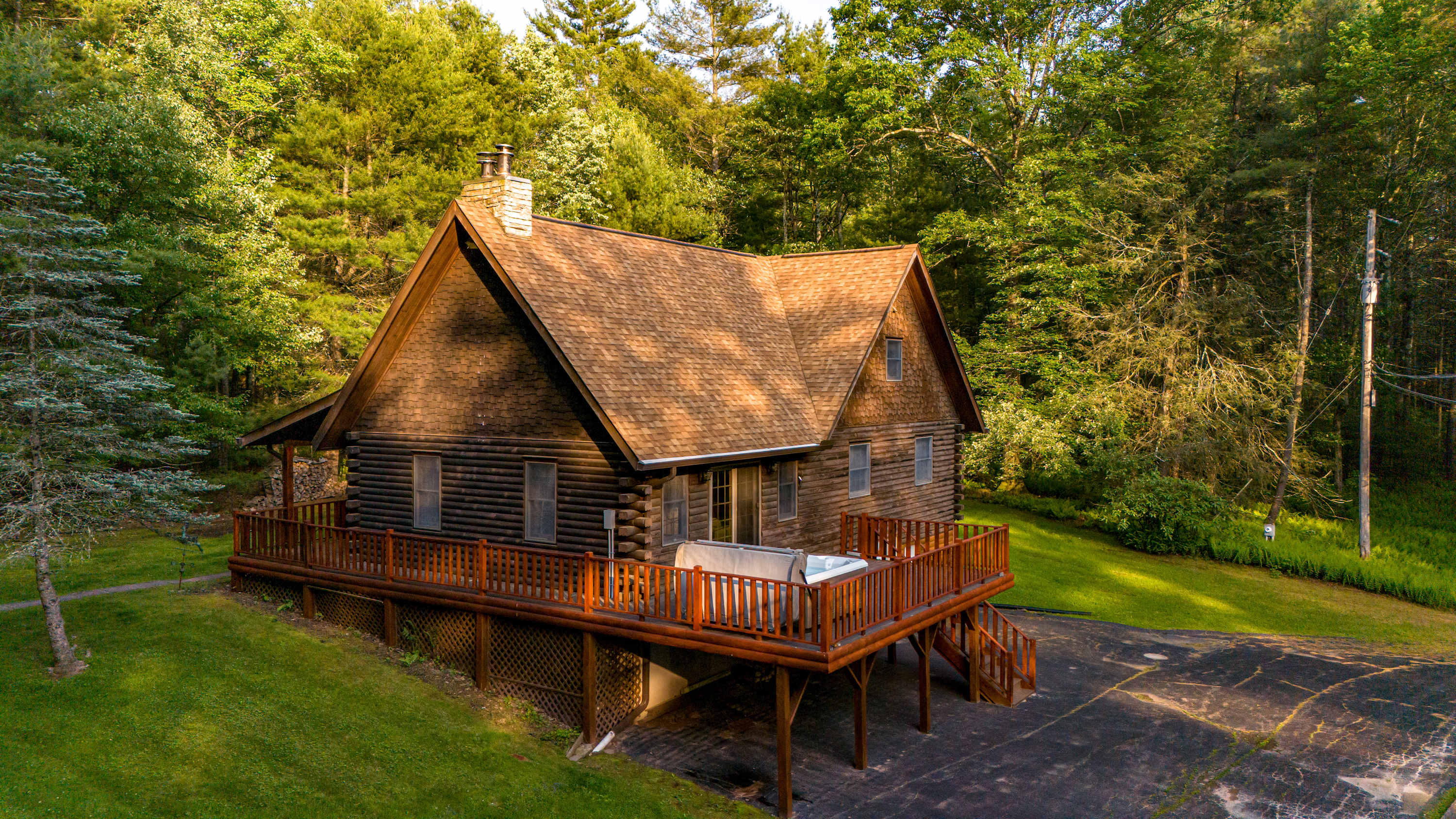 An elevated view of a rustic wooden log cabin with a brown shingle roof, surrounded by dense green forest, showcasing a spacious wooden deck with a hot tub and a paved driveway leading up to the property.