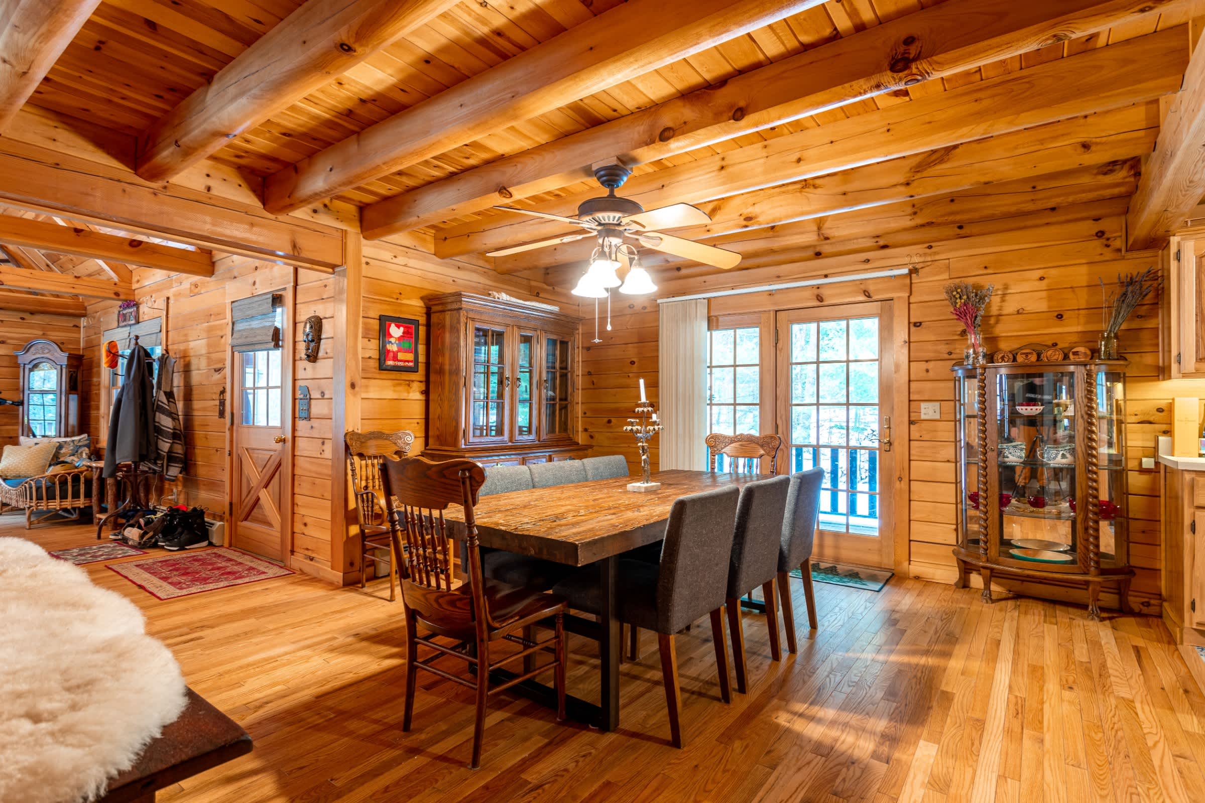 A rustic dining area features a wooden table with upholstered chairs, surrounded by warm wood-paneled walls and a beamed ceiling, with a hutch and a ceiling fan overhead. A glass-paned door leads to an outdoor space, and an open doorway reveals an entryway with a coat rack.
