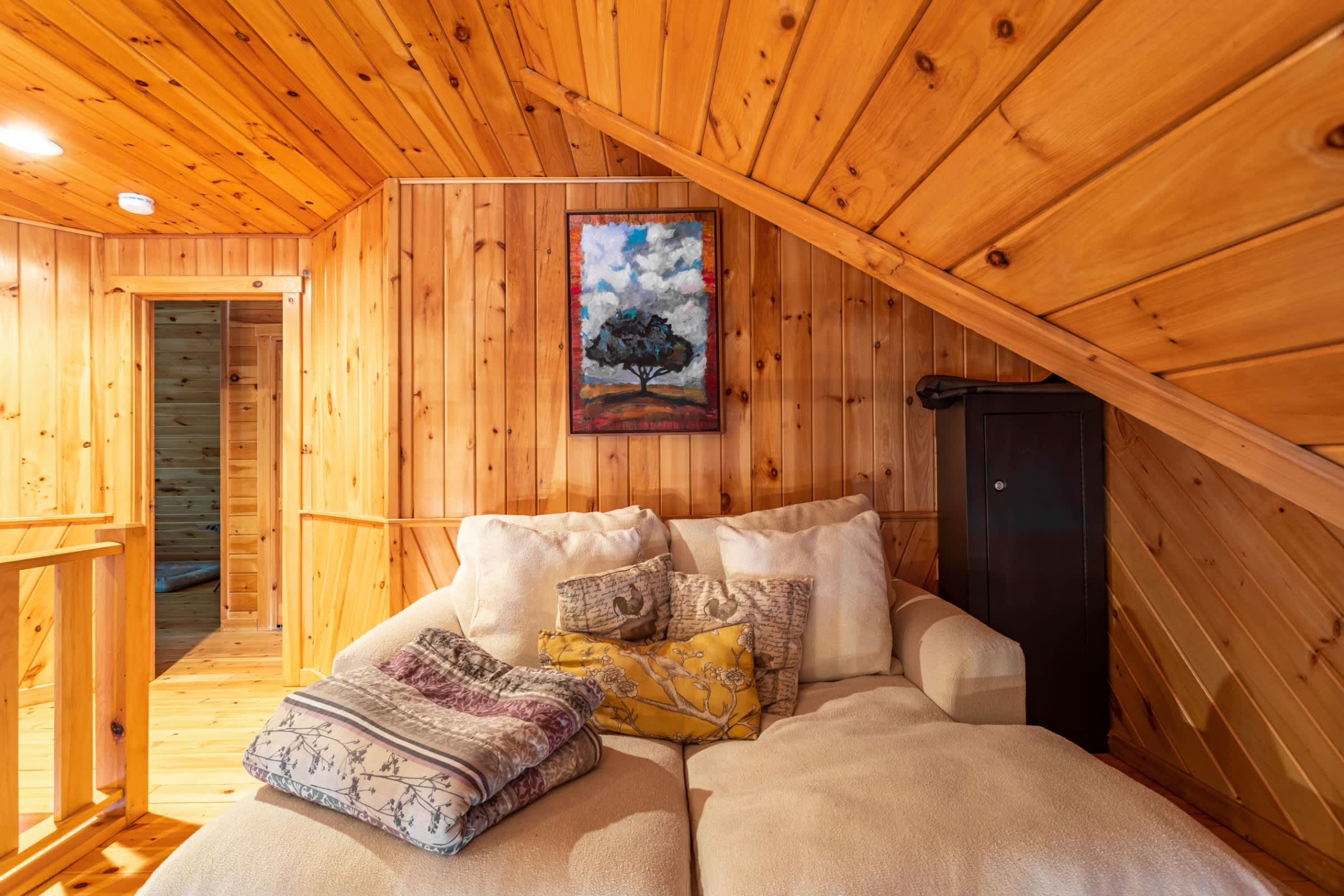 A cozy loft sitting area with warm wooden paneling on the walls and sloped ceiling, furnished with a light-colored chaise lounge adorned with pillows and a blanket, and a colorful abstract painting on the wall.