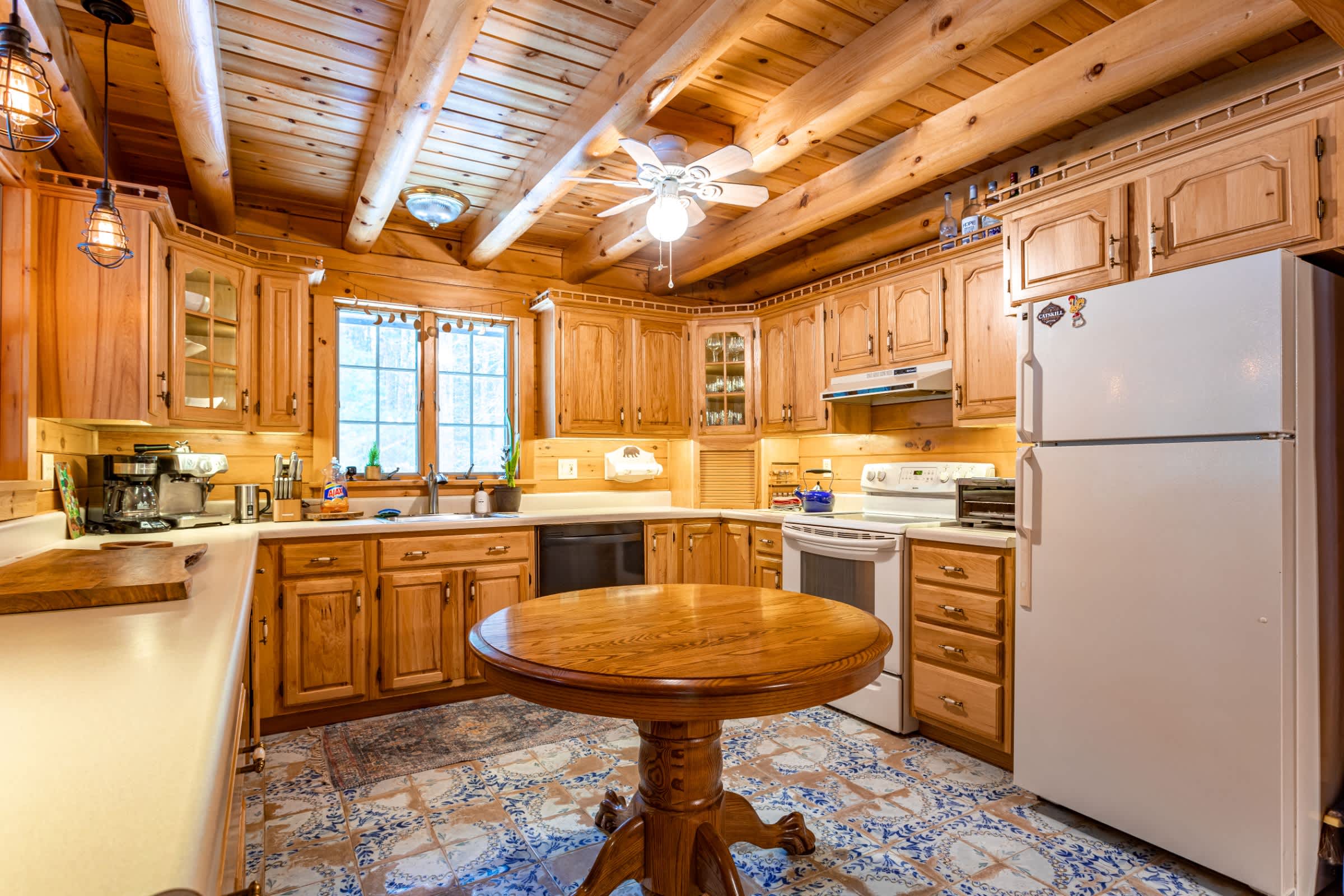 A rustic kitchen with light wood cabinetry, exposed log beams, and a central round wooden table. The floor features blue and white patterned tiles, and white appliances are integrated into the design.