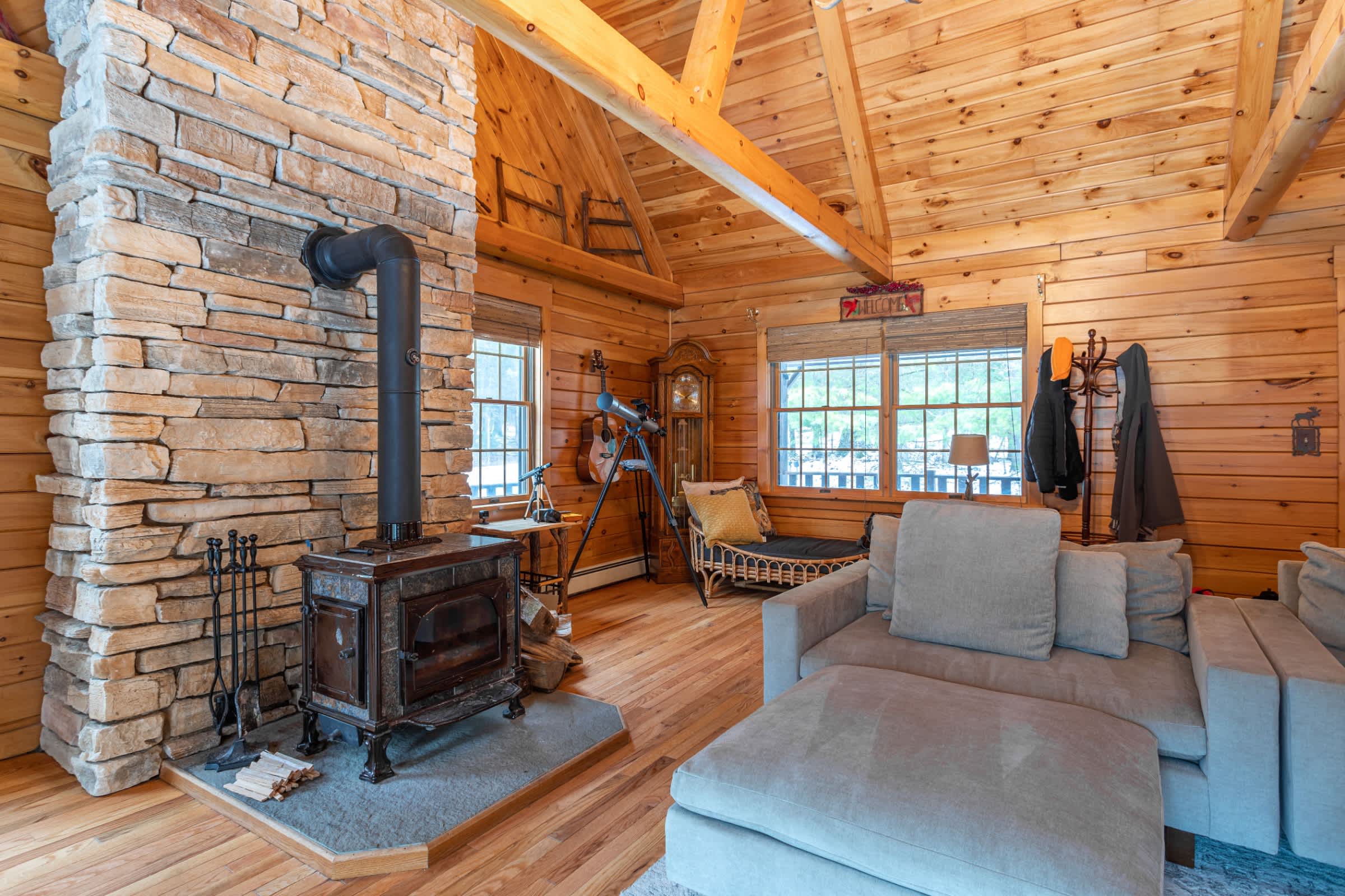 A cozy living room with natural wood walls and vaulted ceilings is centered around a stone fireplace with a black wood-burning stove. A large L-shaped sectional sofa and a telescope are visible.