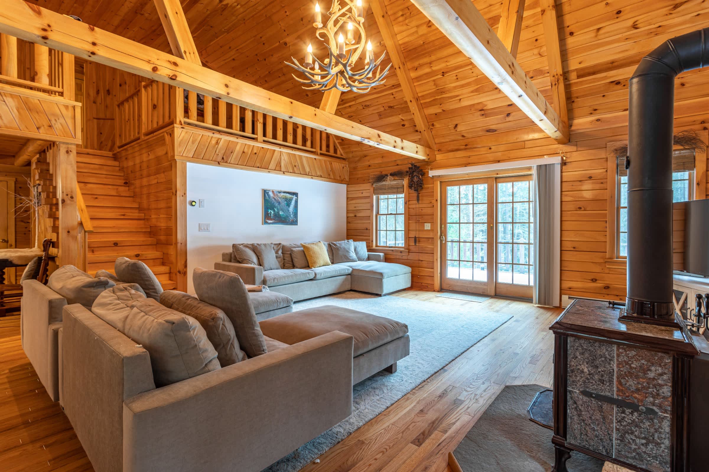 A wide shot of the living room, highlighting the vaulted wood ceiling with exposed beams and a rustic antler chandelier. Two large light gray sectional sofas are arranged on a patterned rug, facing a stone fireplace with a wood-burning stove.