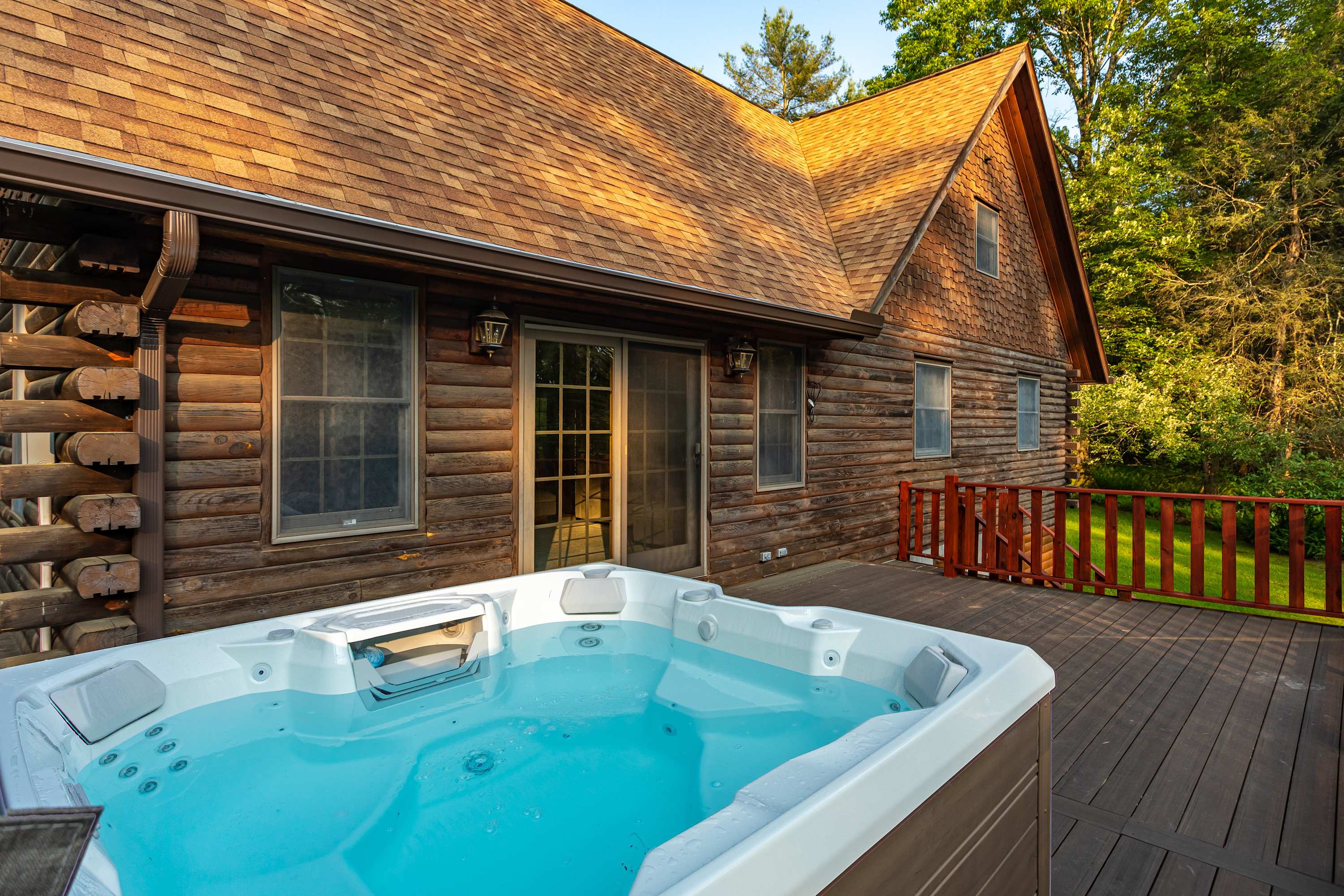 The hot tub is positioned on a wooden deck next to the log cabin exterior, with French doors and windows providing access to the interior. The surrounding area is lush with green trees.