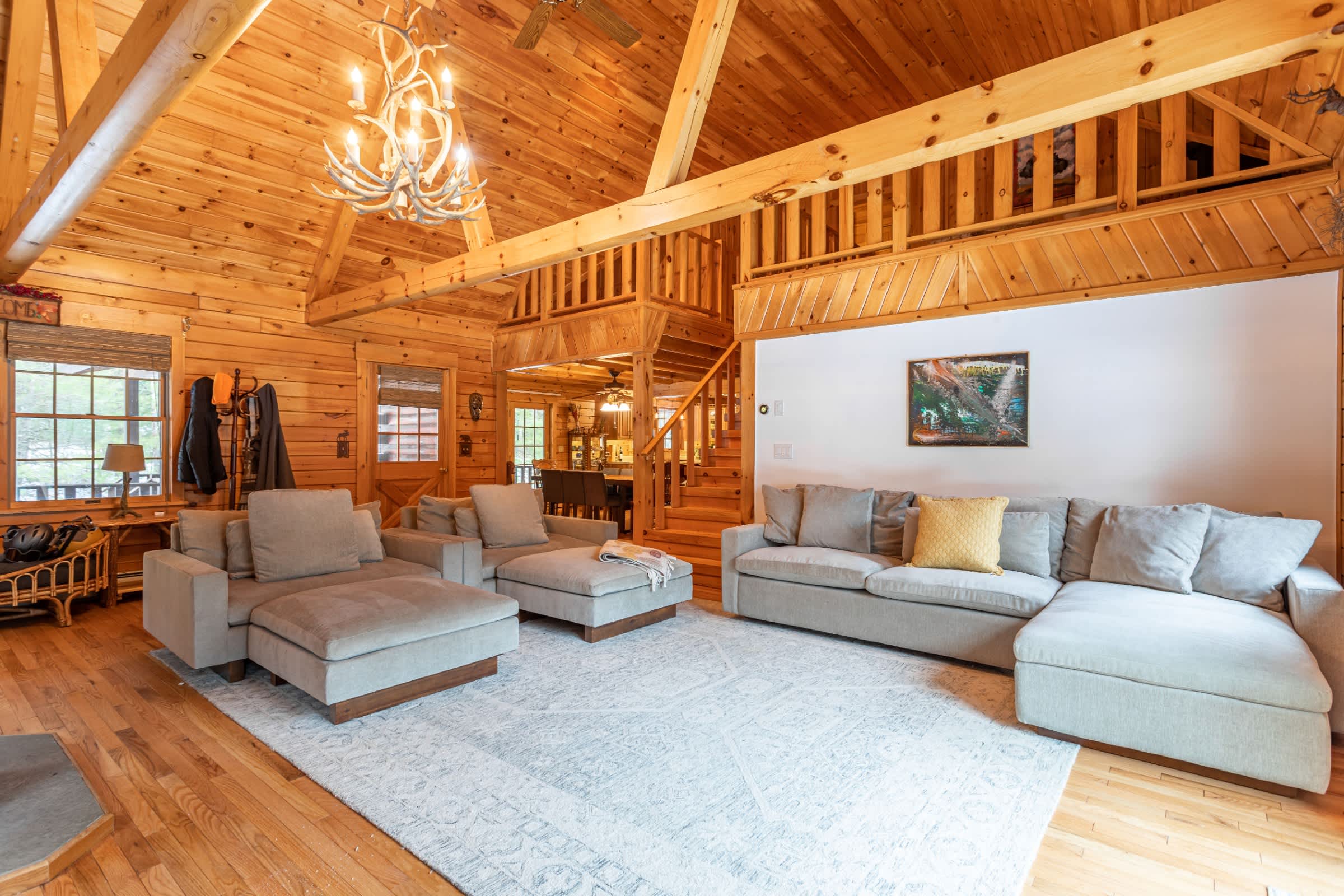 A spacious living room with vaulted wood ceilings and exposed beams, featuring two large light gray sectional sofas on a patterned rug. A rustic antler chandelier hangs from the ceiling, and a wooden staircase leads to an upper level.