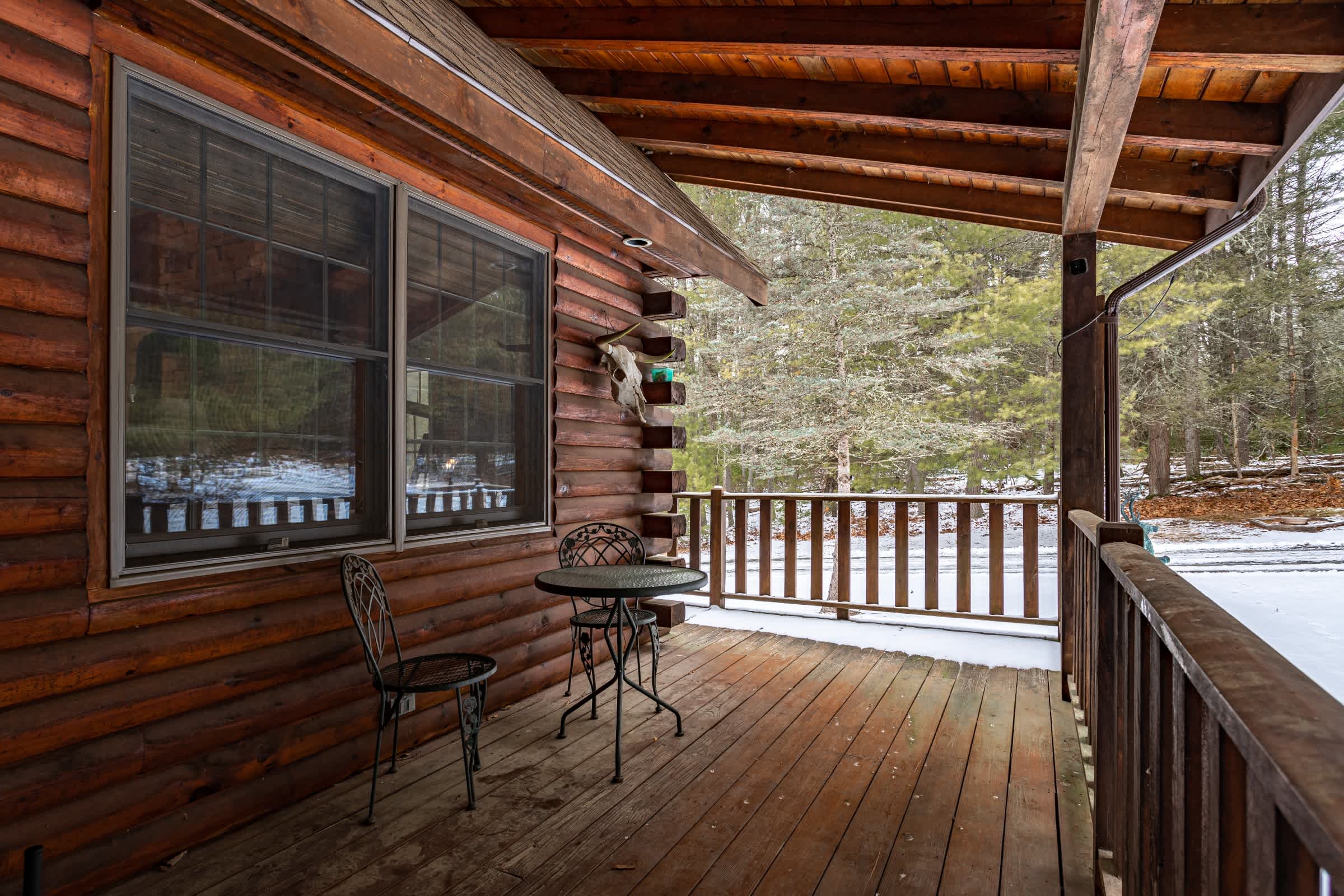 A rustic covered porch with a log cabin exterior, featuring a small round metal table and two chairs on a wooden deck, overlooking a snowy wooded area.