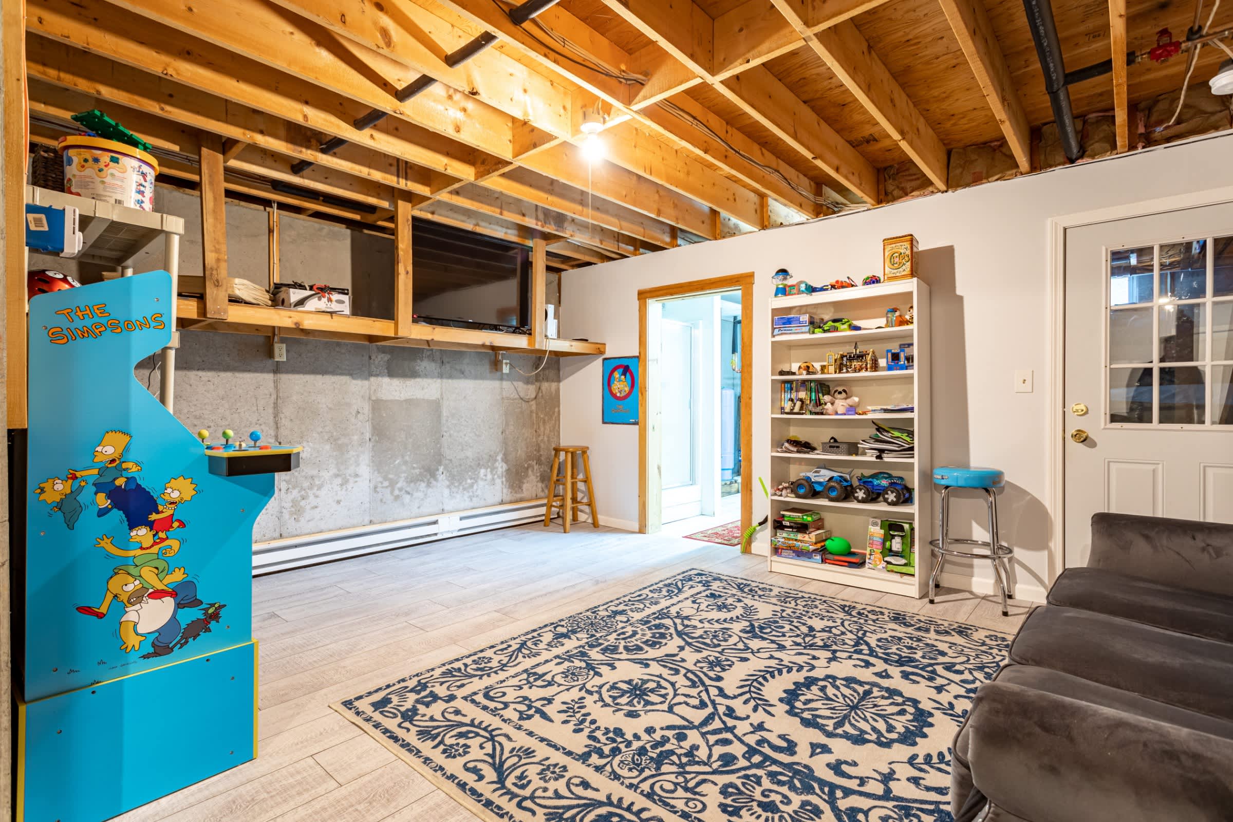 A basement game room features exposed ceiling joists, light wood-look flooring, and concrete walls. It includes a Simpsons arcade machine, a wall-mounted TV, and a white bookshelf with toys.