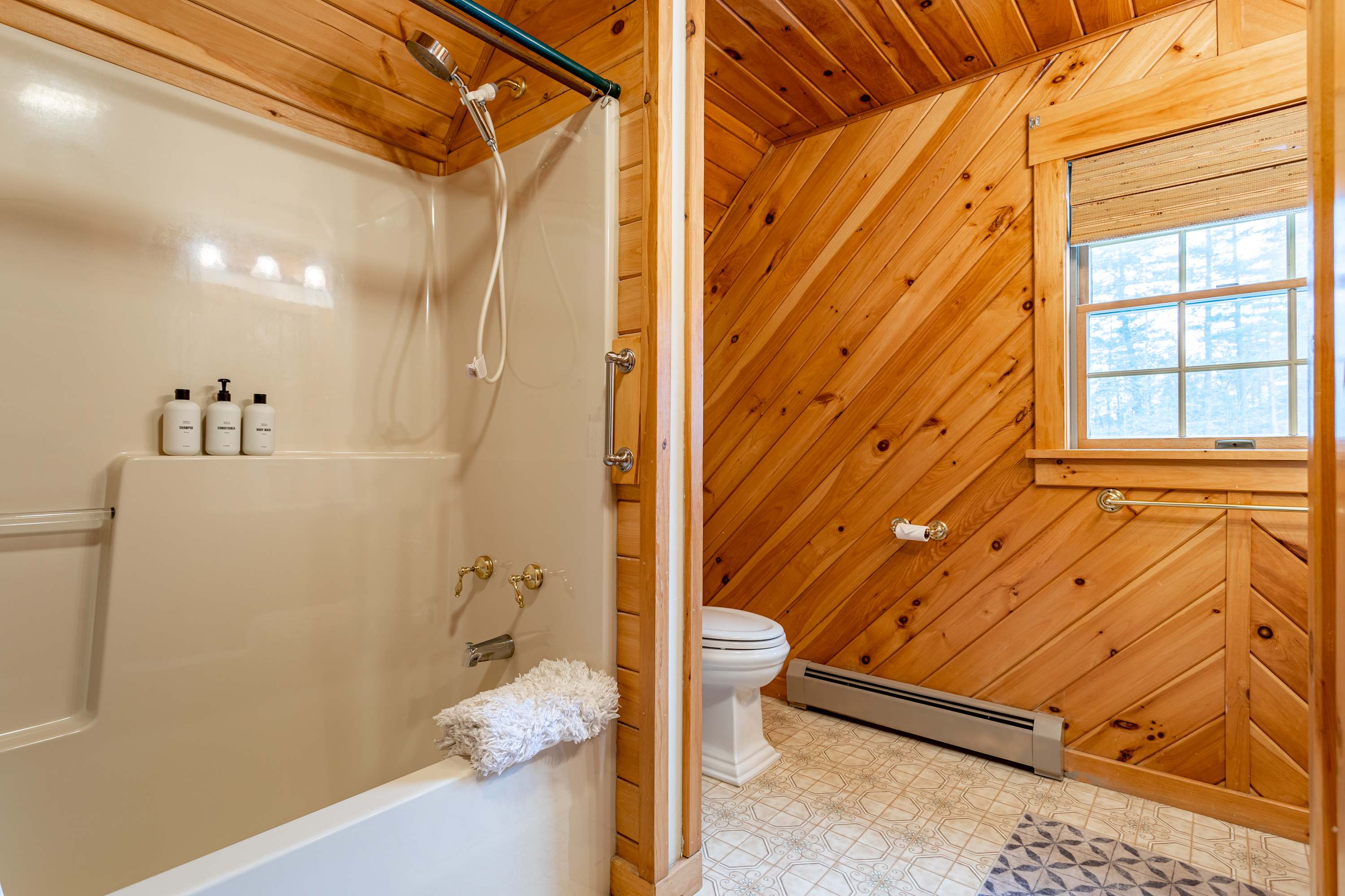 The rustic bathroom continues with wood-paneled walls and a patterned floor, featuring a white shower-tub combination with a showerhead and faucet. A small window is visible above the toilet, which is next to the shower.