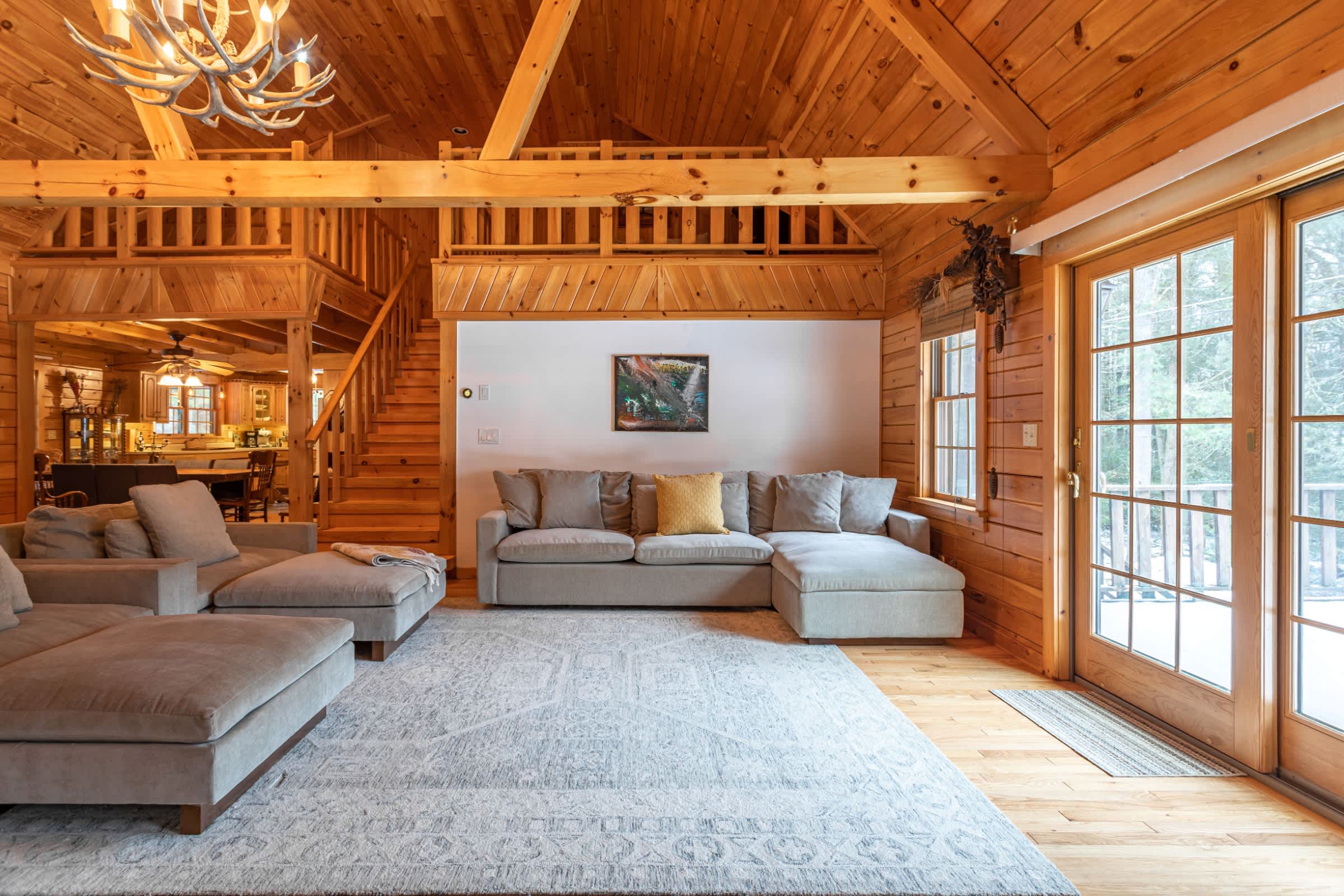 A spacious living room features a large L-shaped sectional sofa on a light patterned rug, set against a white accent wall with a painting. The room boasts warm wood-paneled walls, a high beamed ceiling, and large glass doors opening to the exterior, with a wooden staircase leading to an upper level.