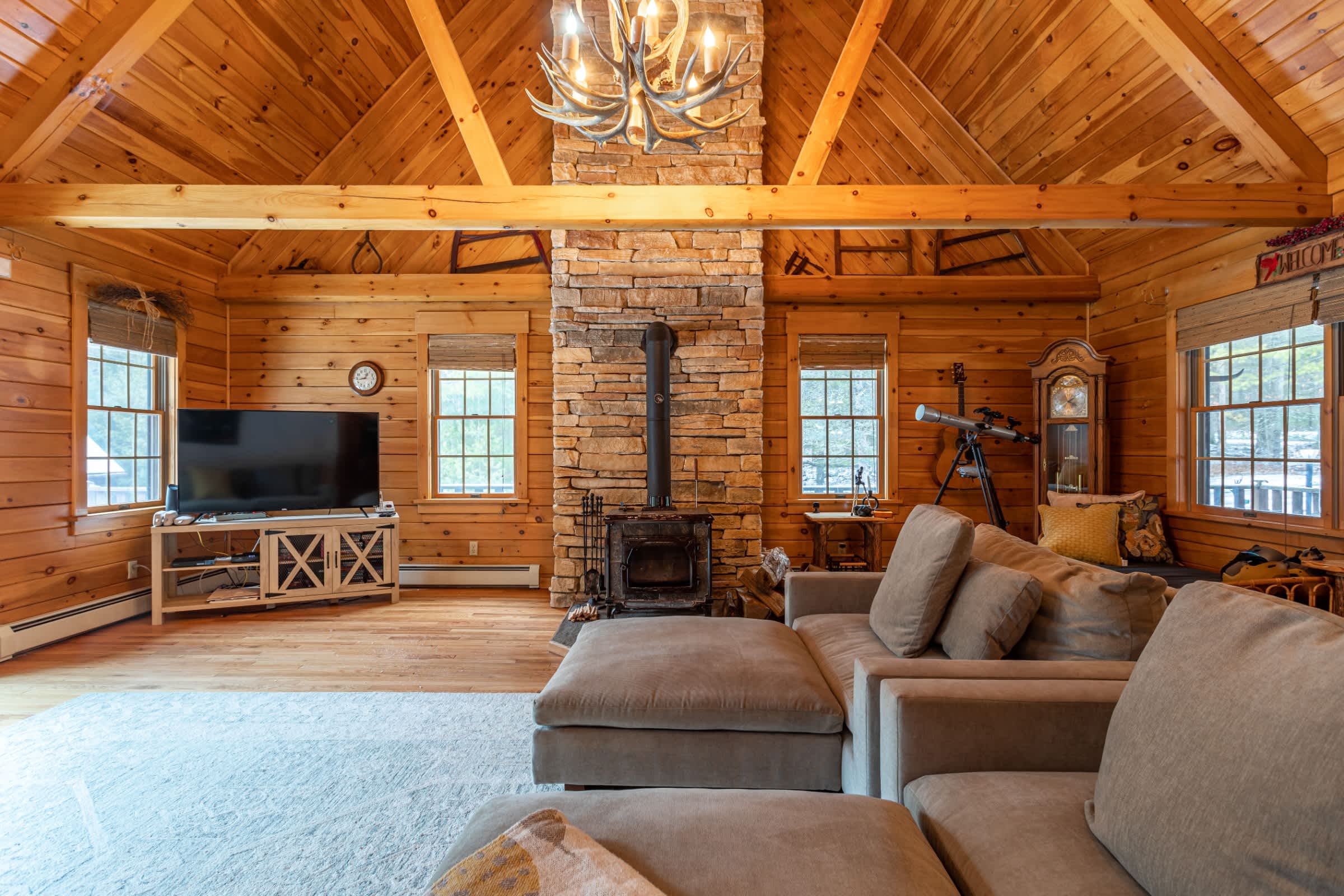 A spacious living room with natural wood walls and a high vaulted ceiling with exposed beams is centered around a stone fireplace. A large sectional sofa, TV, and antler chandelier complete the rustic decor.