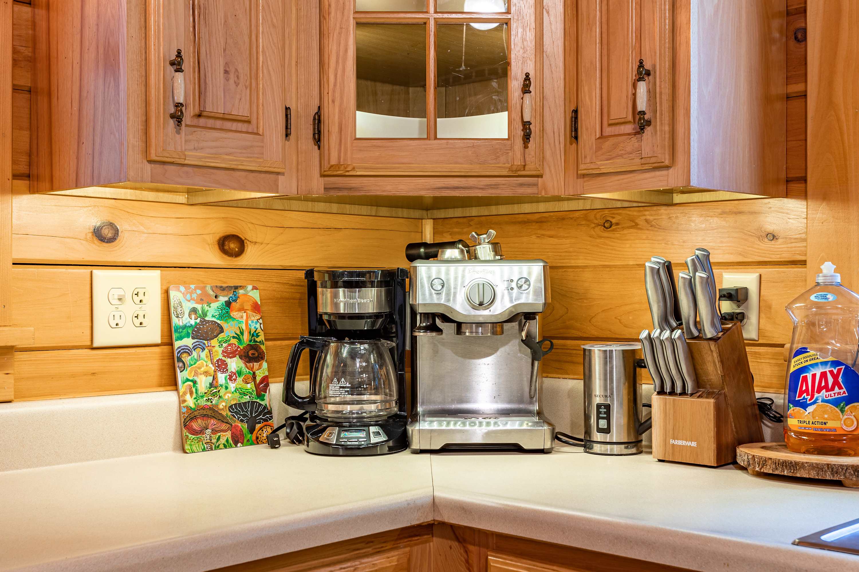 A rustic kitchen view shows light wood cabinetry, white appliances, and a round wooden table in the foreground. Beyond the kitchen, a dining area with a long wooden table and chairs is visible.
