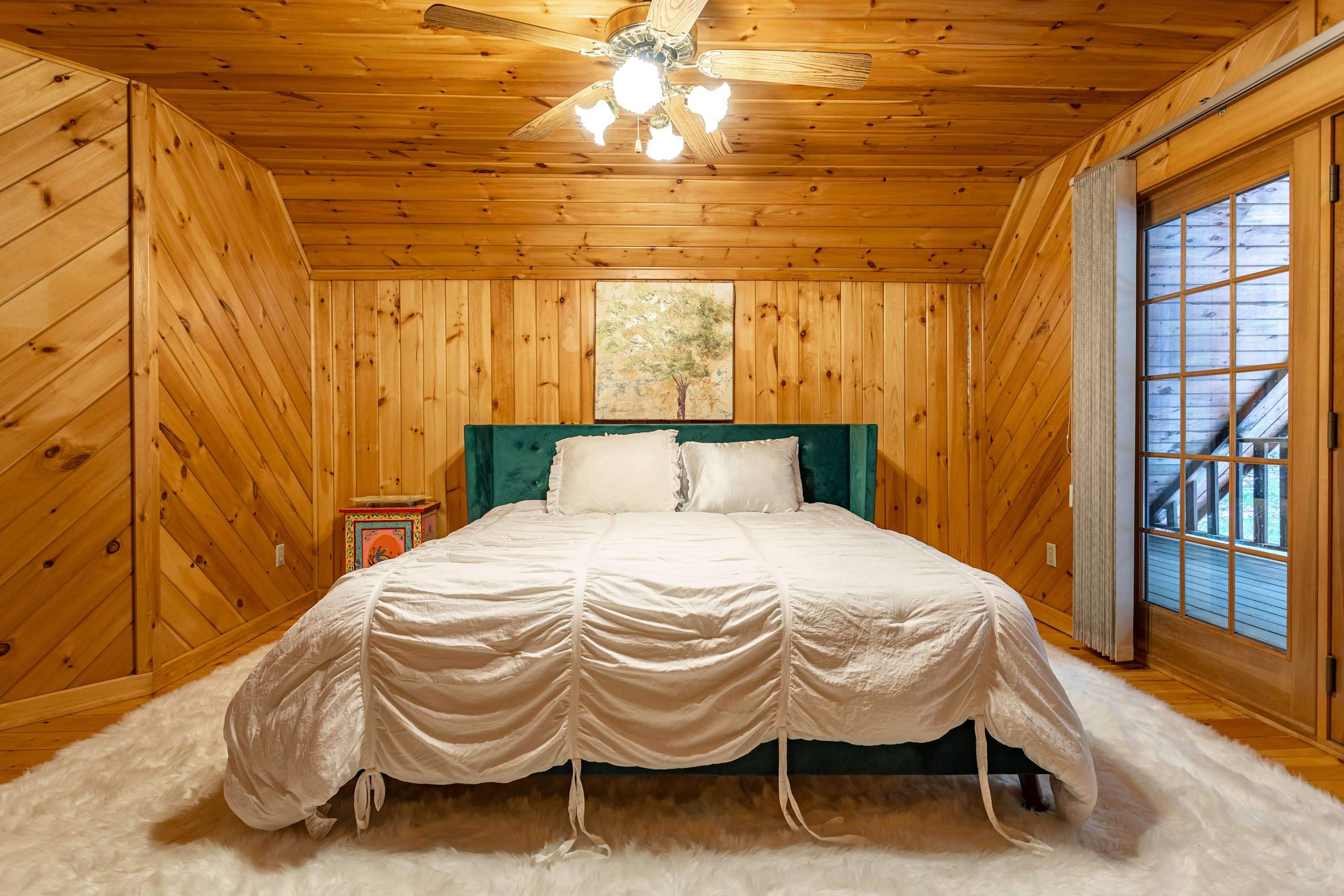 A cozy bedroom features a large bed with a white ruffled duvet and a green velvet headboard, centered on a plush white rug. The room is enveloped in warm wood-paneled walls and a matching sloped ceiling, with a ceiling fan and a glass-paned door leading to an outdoor area.