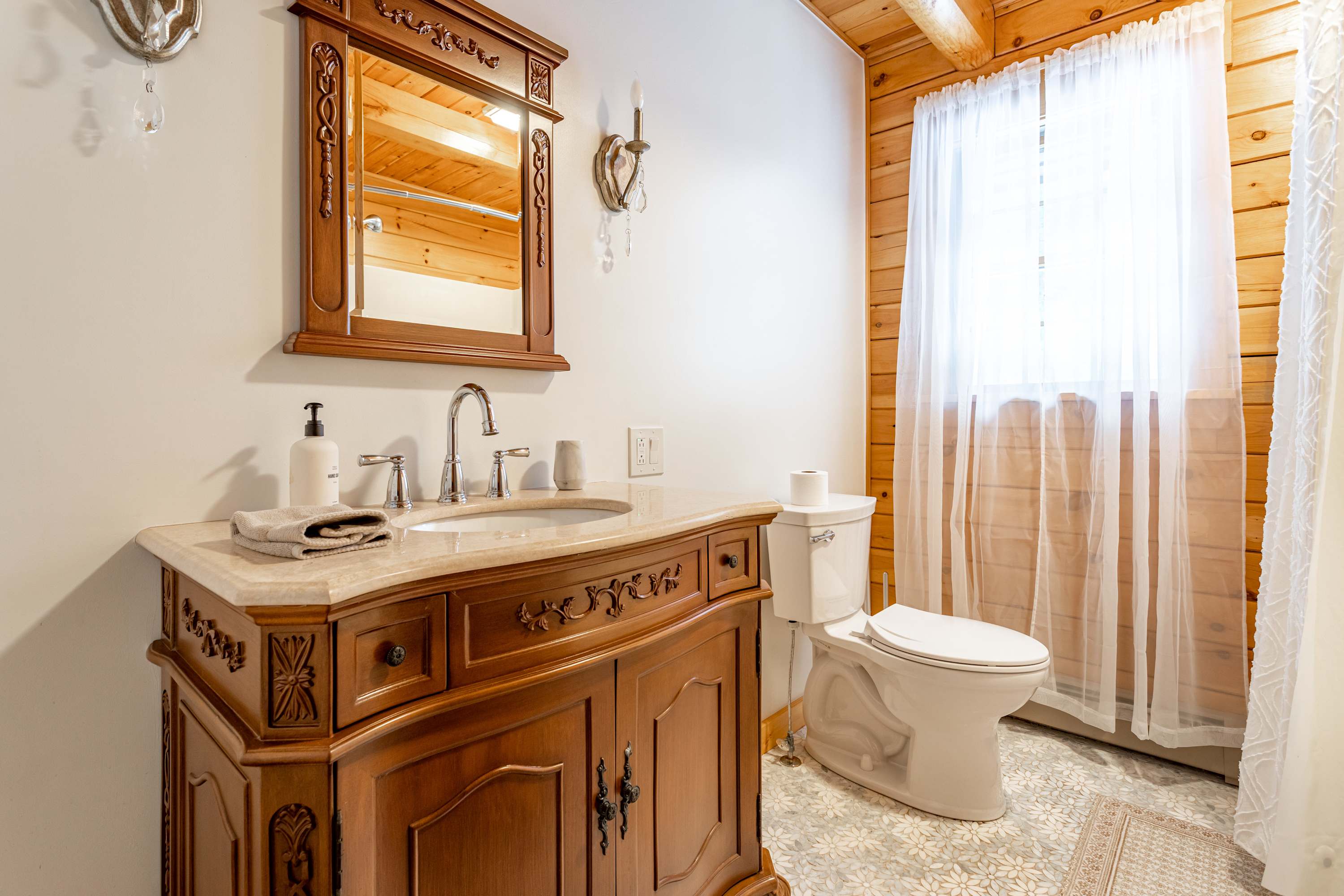 This elegant bathroom showcases a carved wooden vanity with a marble top and sink, paired with an ornate mirror and sconces. A white toilet is positioned next to a window draped with sheer white curtains, all set against white walls and a wood-beamed ceiling.