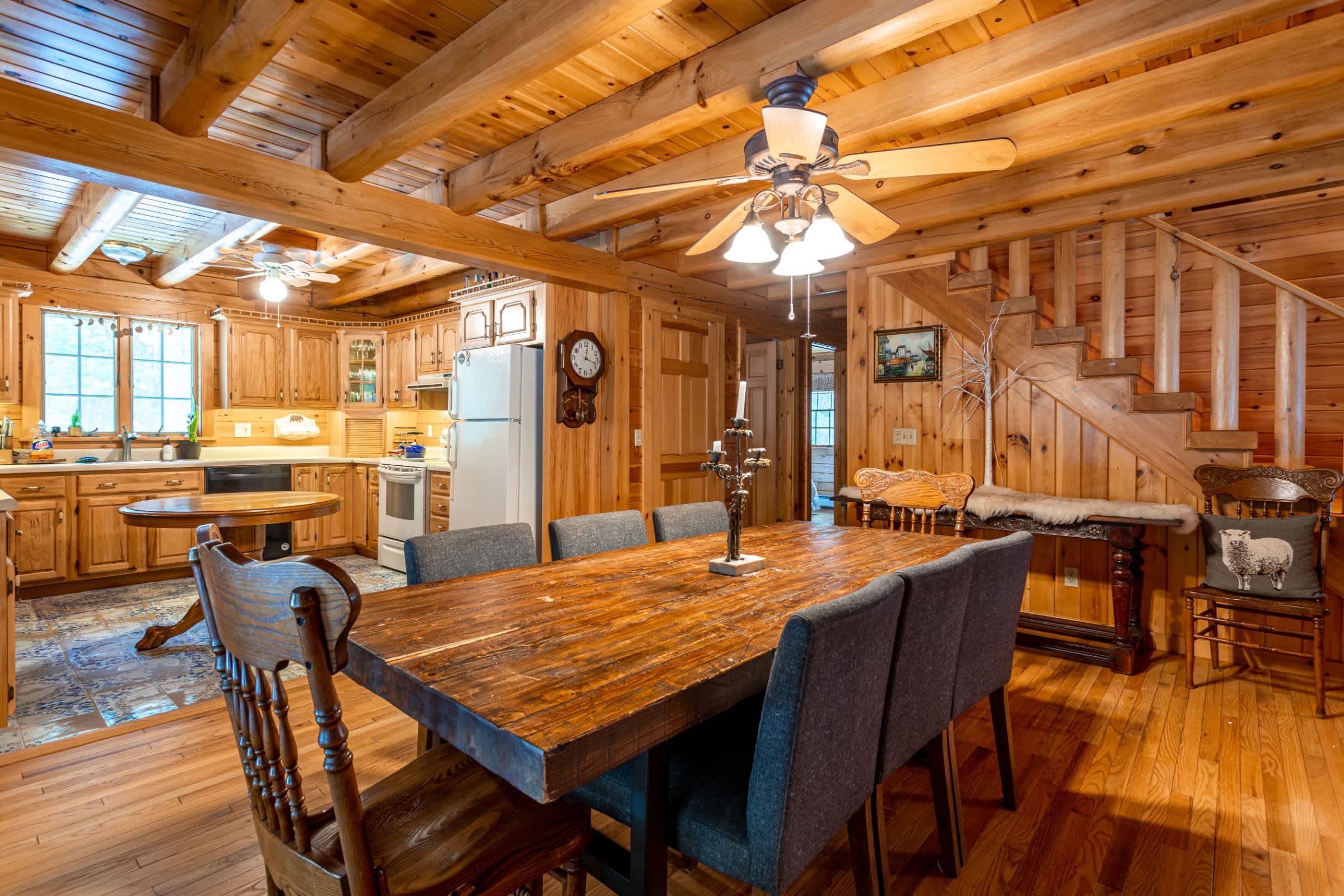 A rustic dining area with a large wooden table and upholstered chairs, adjacent to an open-concept kitchen with light wood cabinetry and white appliances. The room features wood walls, ceiling, and flooring, with a wooden staircase in the background.