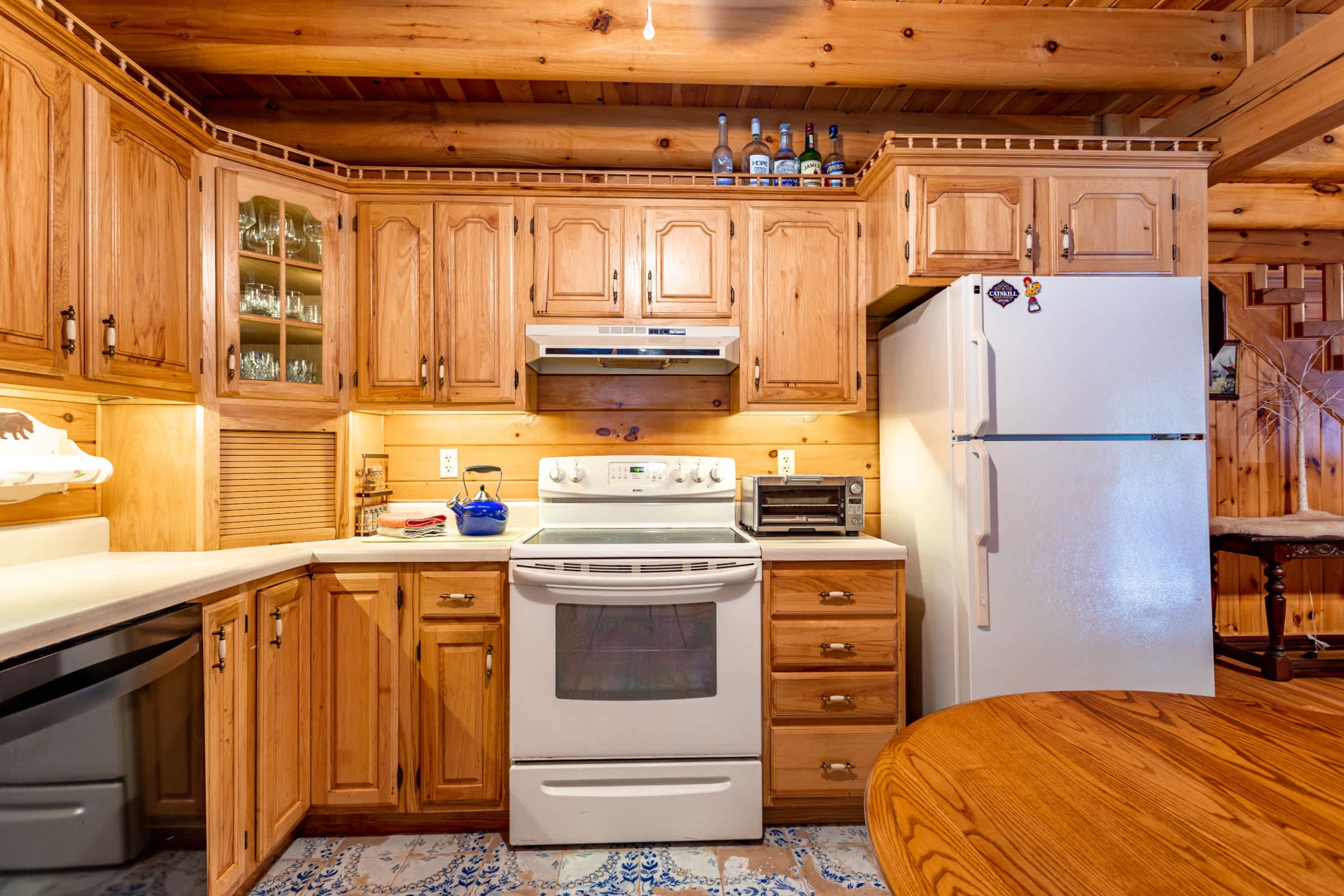 A close-up view of the rustic kitchen highlights light wood cabinetry, white appliances including a stove and refrigerator, and the blue and white patterned tile floor. A round wooden table is in the foreground.