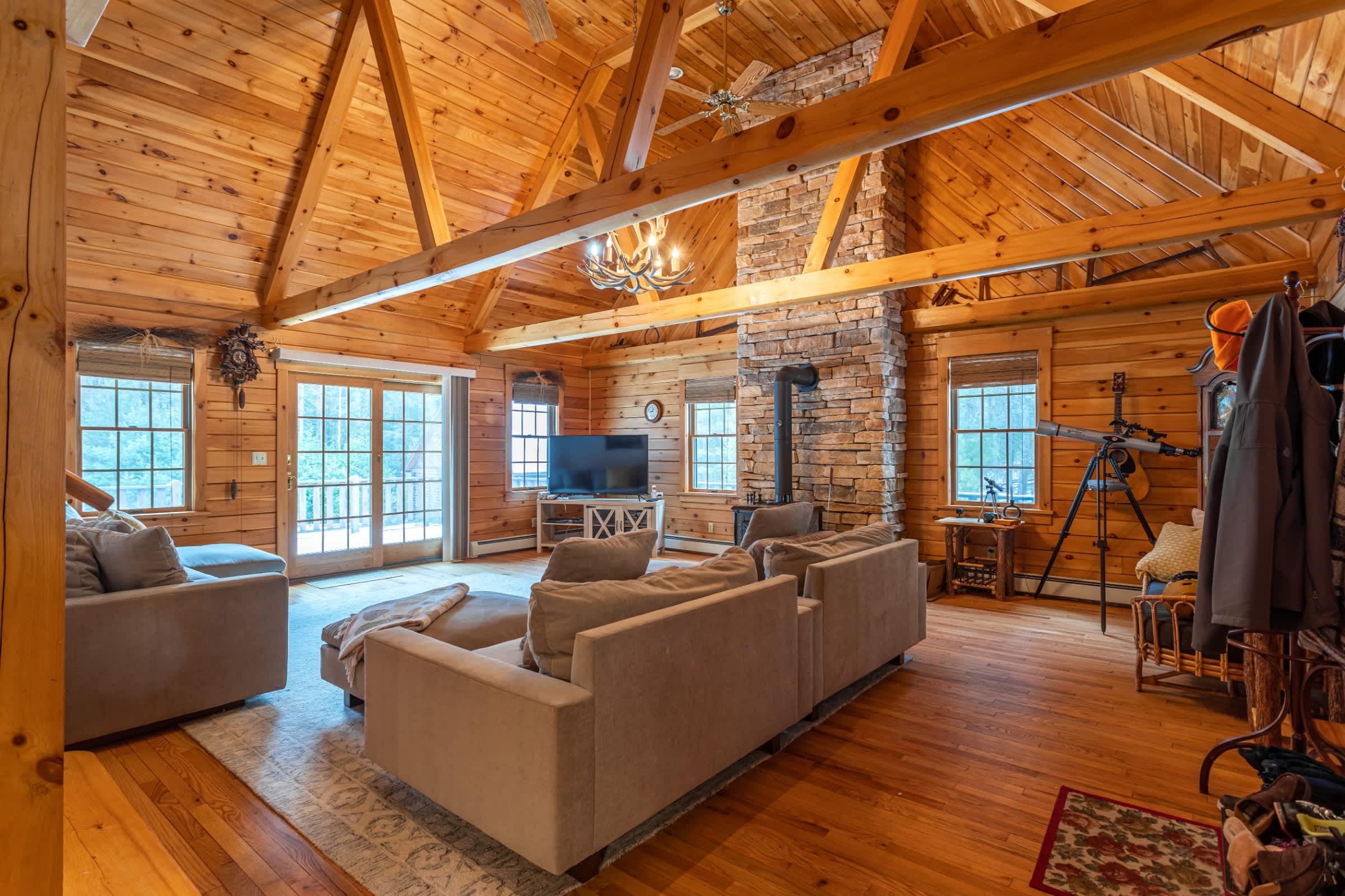 A wide view of the living room showcasing the vaulted wood ceilings, exposed beams, and a large stone fireplace with a wood-burning stove. Multiple light gray sofas are arranged around a patterned rug, with French doors leading to the outside.