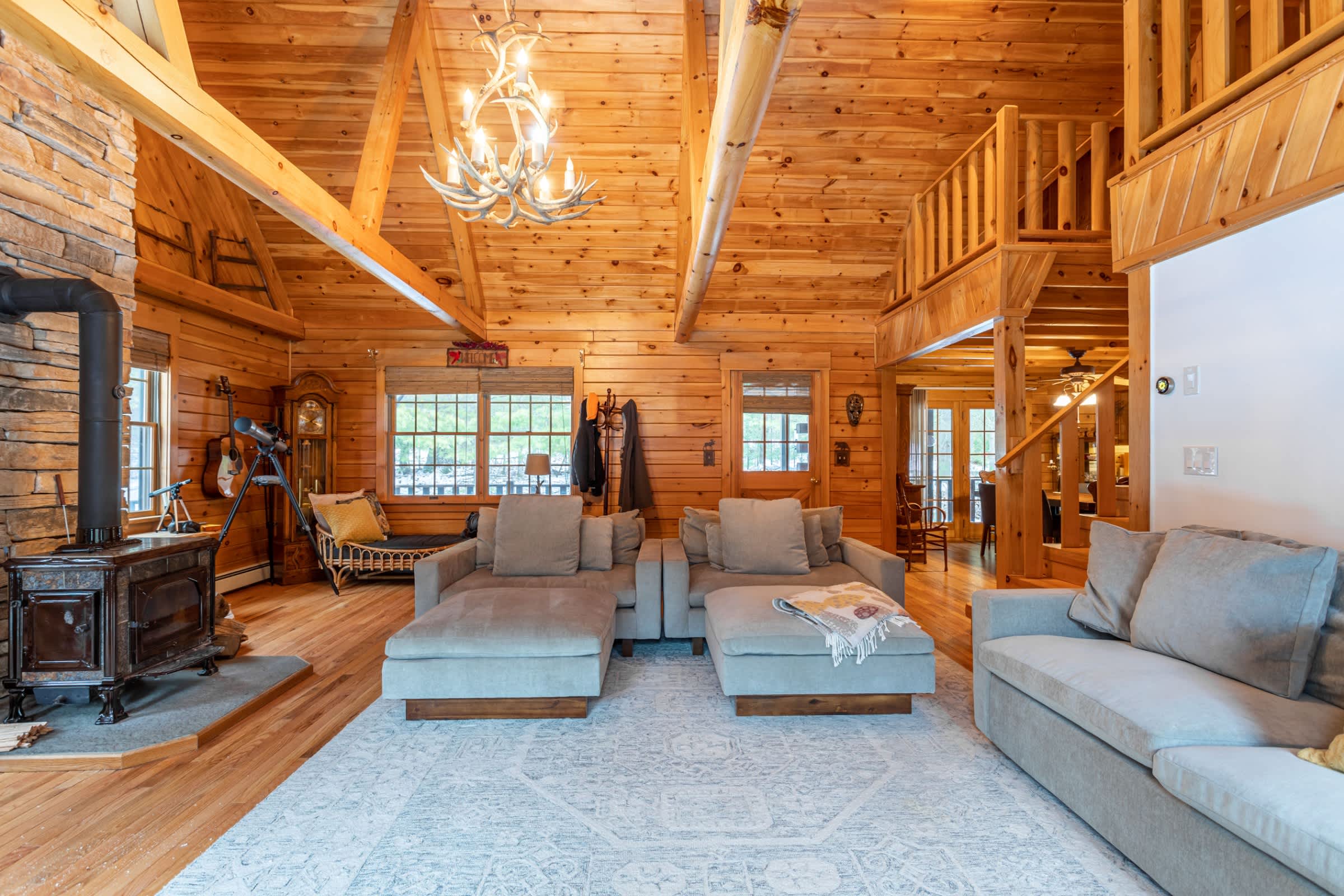 A wide view of the rustic living room features natural wood walls, vaulted ceilings, and a large L-shaped sectional sofa on a light rug. A stone fireplace and a wooden staircase leading upstairs are visible.