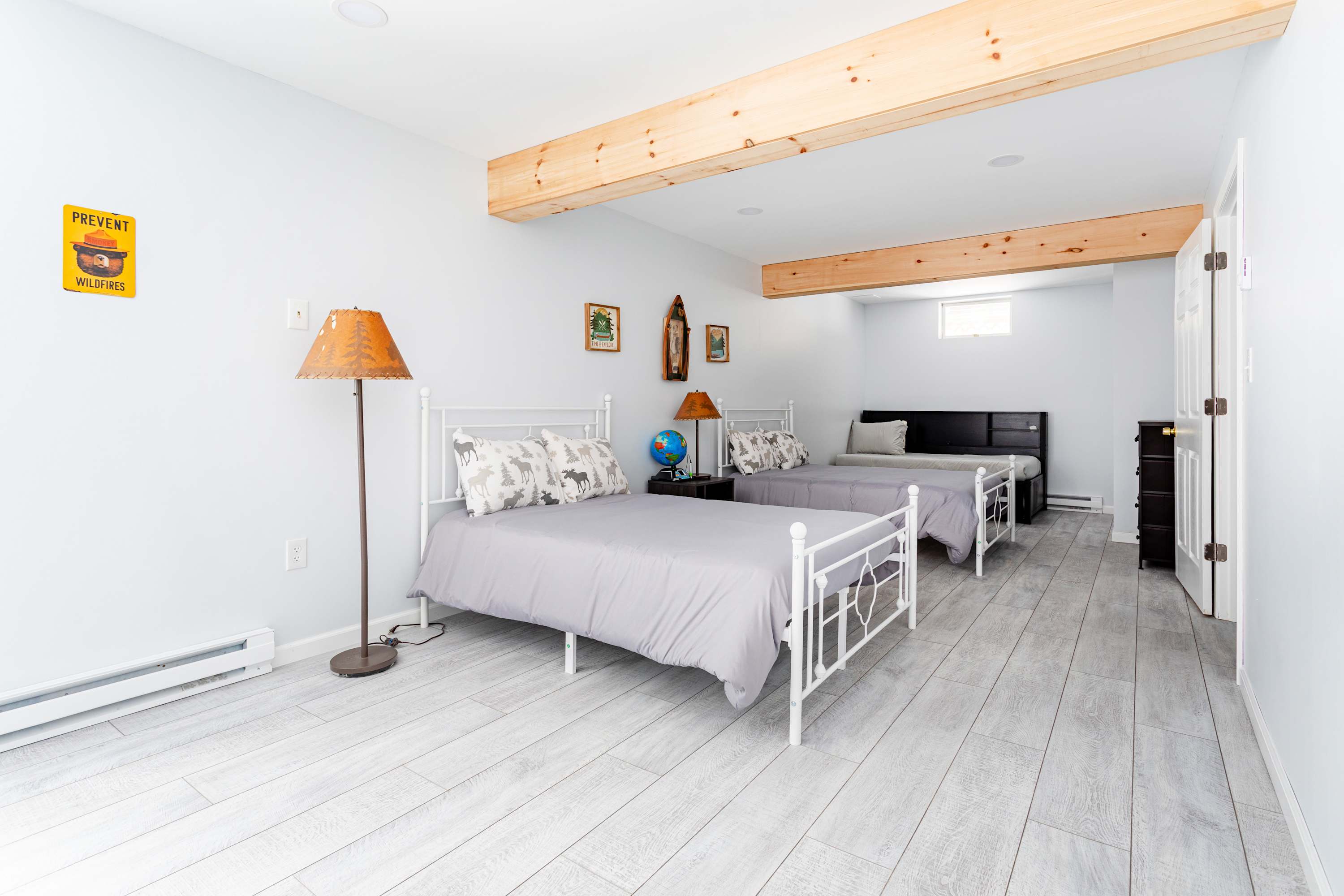 A bright bedroom featuring two white metal-frame twin beds with grey bedding, light wood-look flooring, and exposed wooden ceiling beams, with a third bed visible in the background.