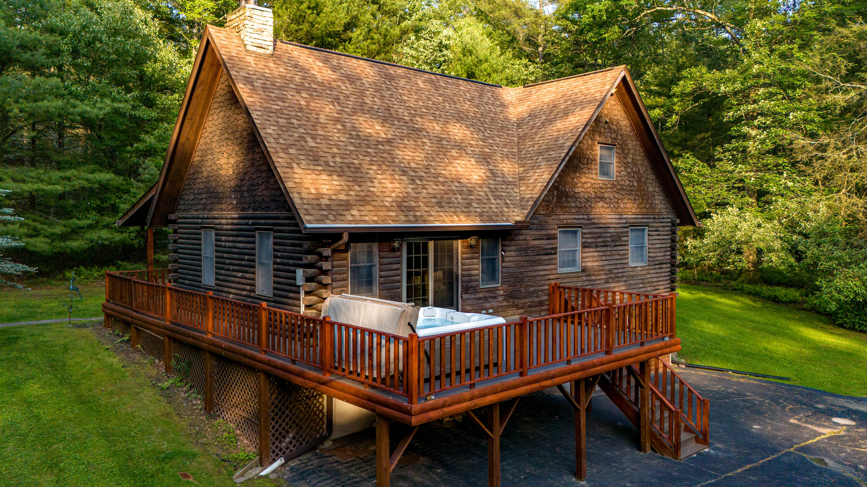 An aerial view of a charming wooden log cabin nestled among lush green trees, featuring a brown shingle roof, a large wraparound wooden deck, and a hot tub, with a paved driveway in the foreground.