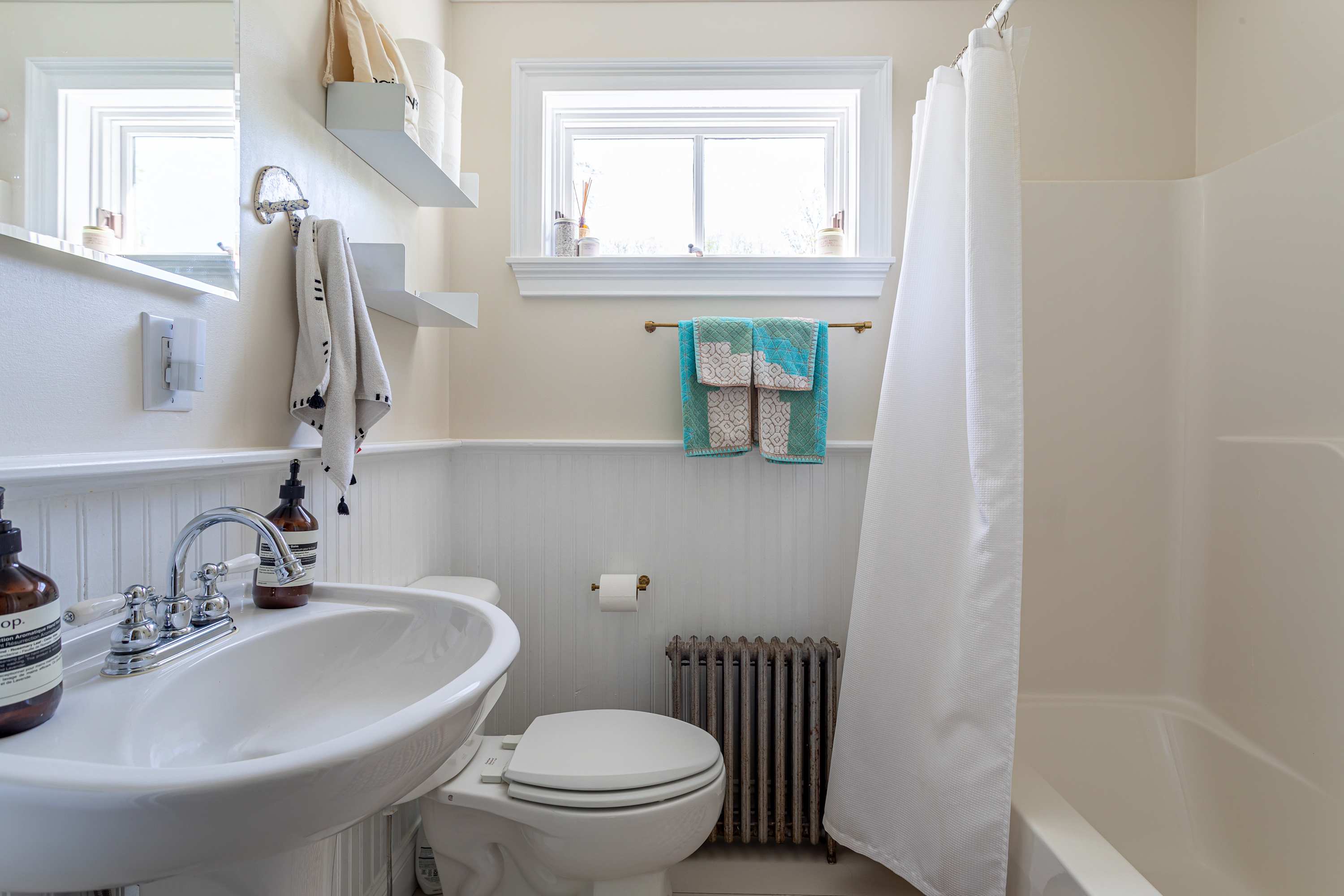 A light-filled full bathroom featuring a white pedestal sink, a toilet, and a shower/tub combo with a white curtain. White wainscoting lines the walls, and a window is positioned above the toilet.