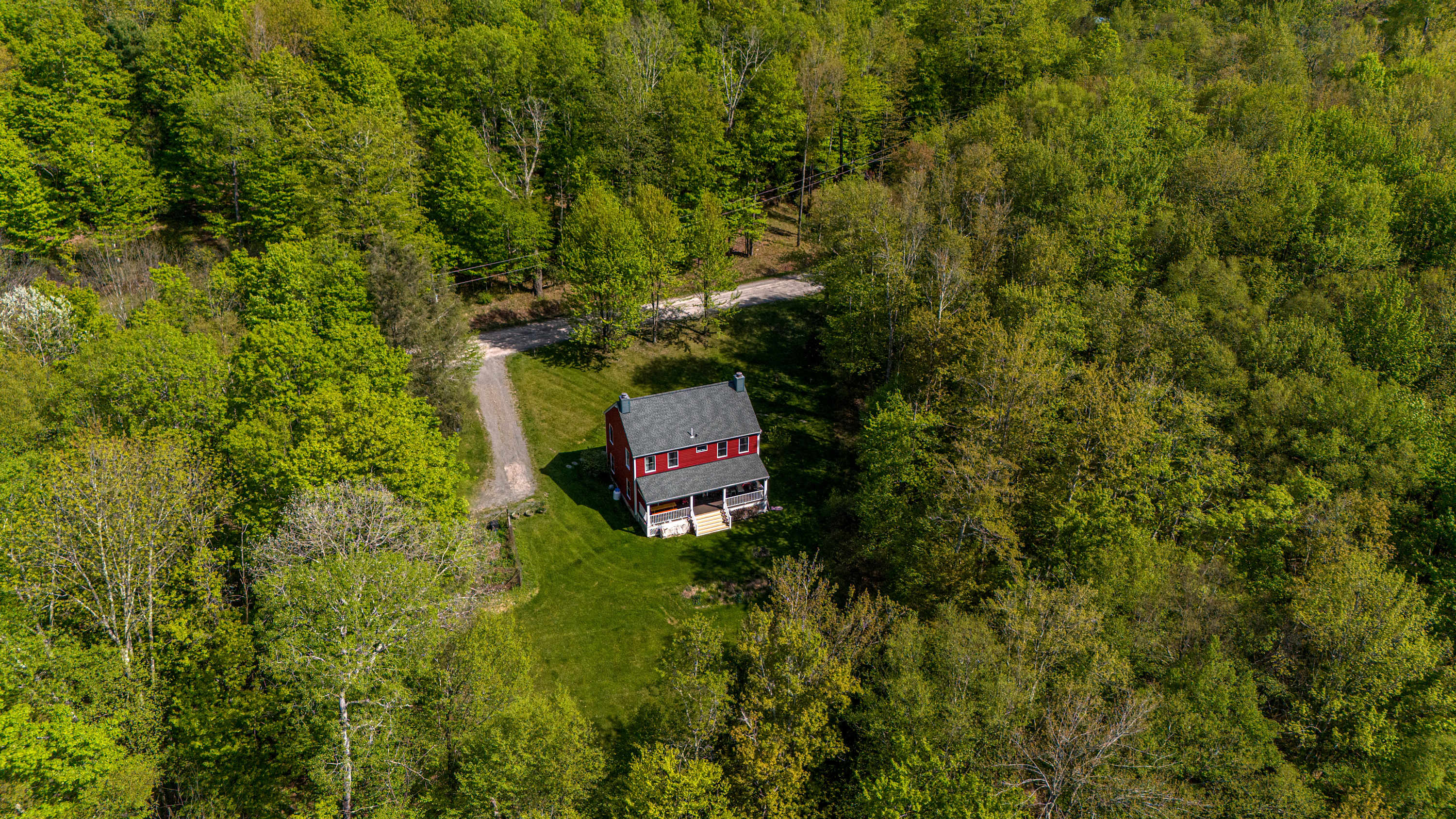 Stunning aerial view of the red farmhouse nestled amidst a dense, vibrant green forest, with a winding dirt driveway leading to the property. The house stands out against the natural landscape, showcasing its secluded and private setting.