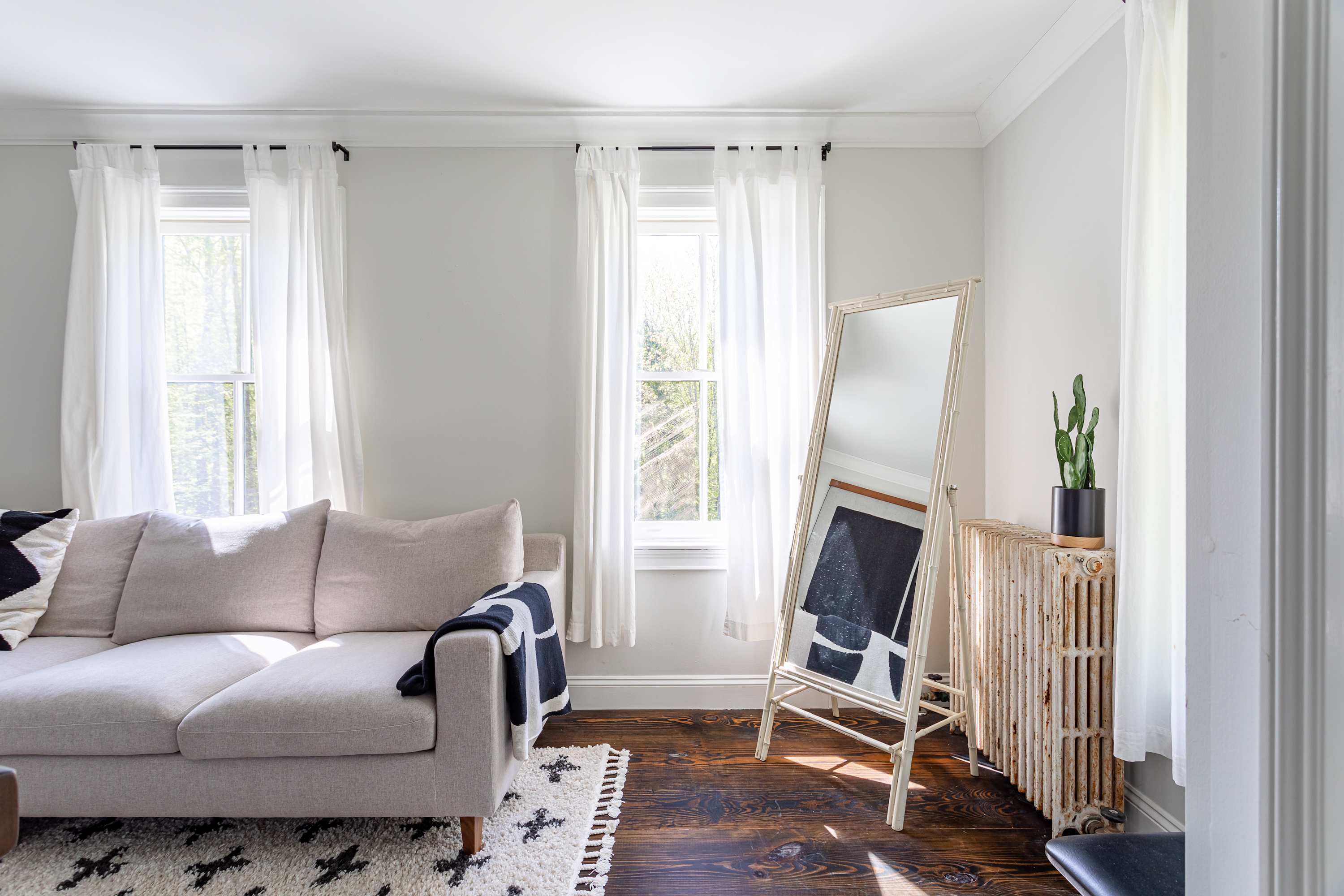 Bright living room featuring a light-colored sofa with a black and white throw, dark hardwood floors, and two large windows with white sheer curtains. A tall, ornate mirror leans against a white radiator, reflecting a dark abstract artwork.