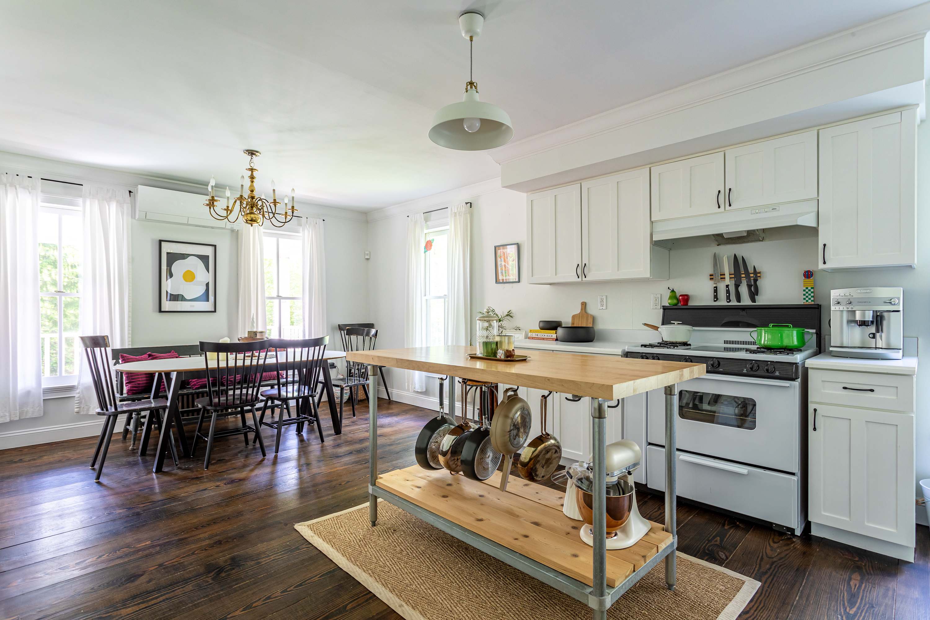 Spacious open-concept kitchen and dining area with white shaker cabinets, a butcher block island, and a long white dining table with black chairs and a bench. Dark hardwood floors and white walls create a bright space.