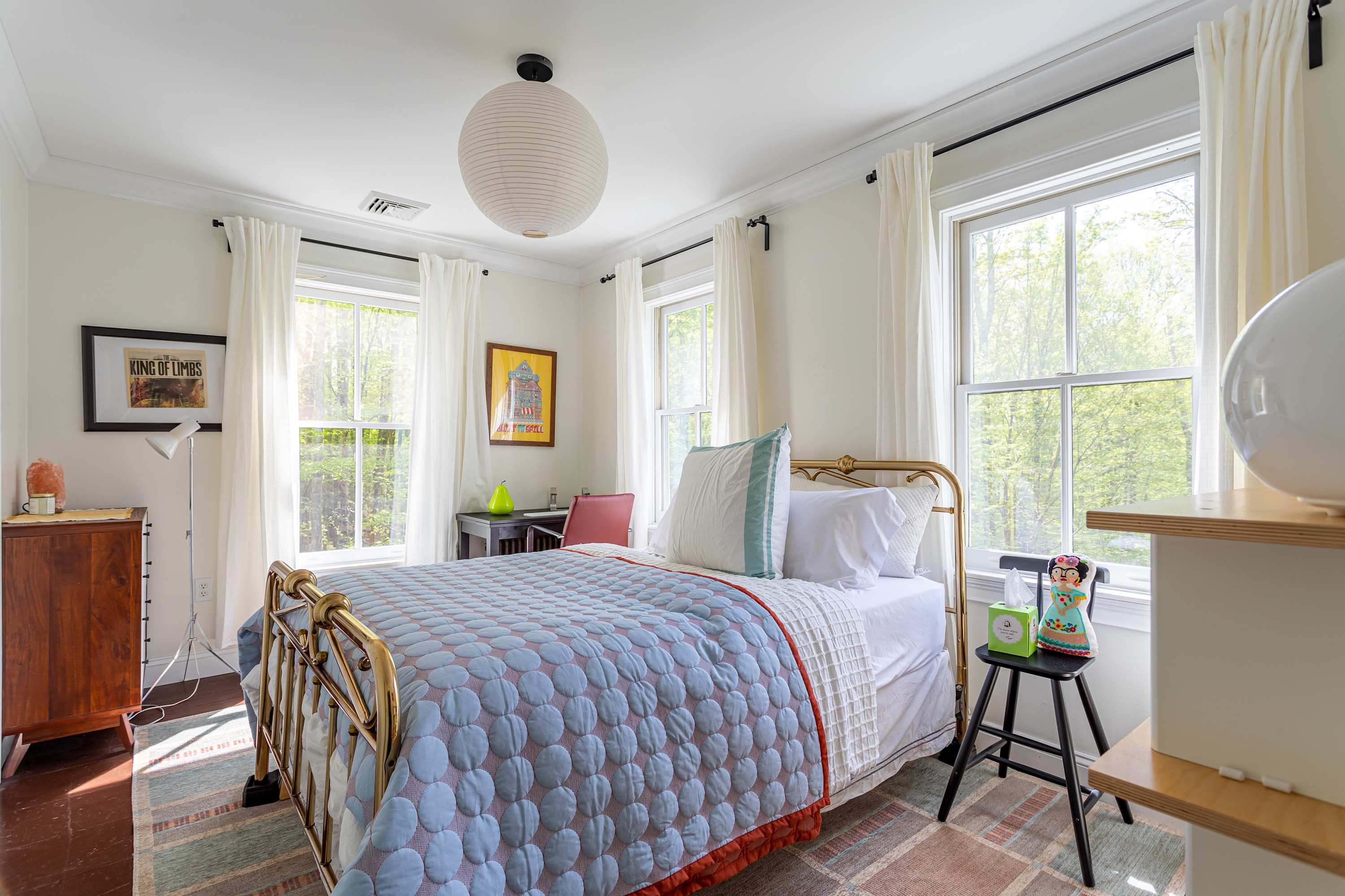 A bright bedroom featuring a brass bed frame with a blue and white quilted comforter, flanked by large windows with white curtains. A white paper lantern hangs from the ceiling, and a small desk is visible in the corner.