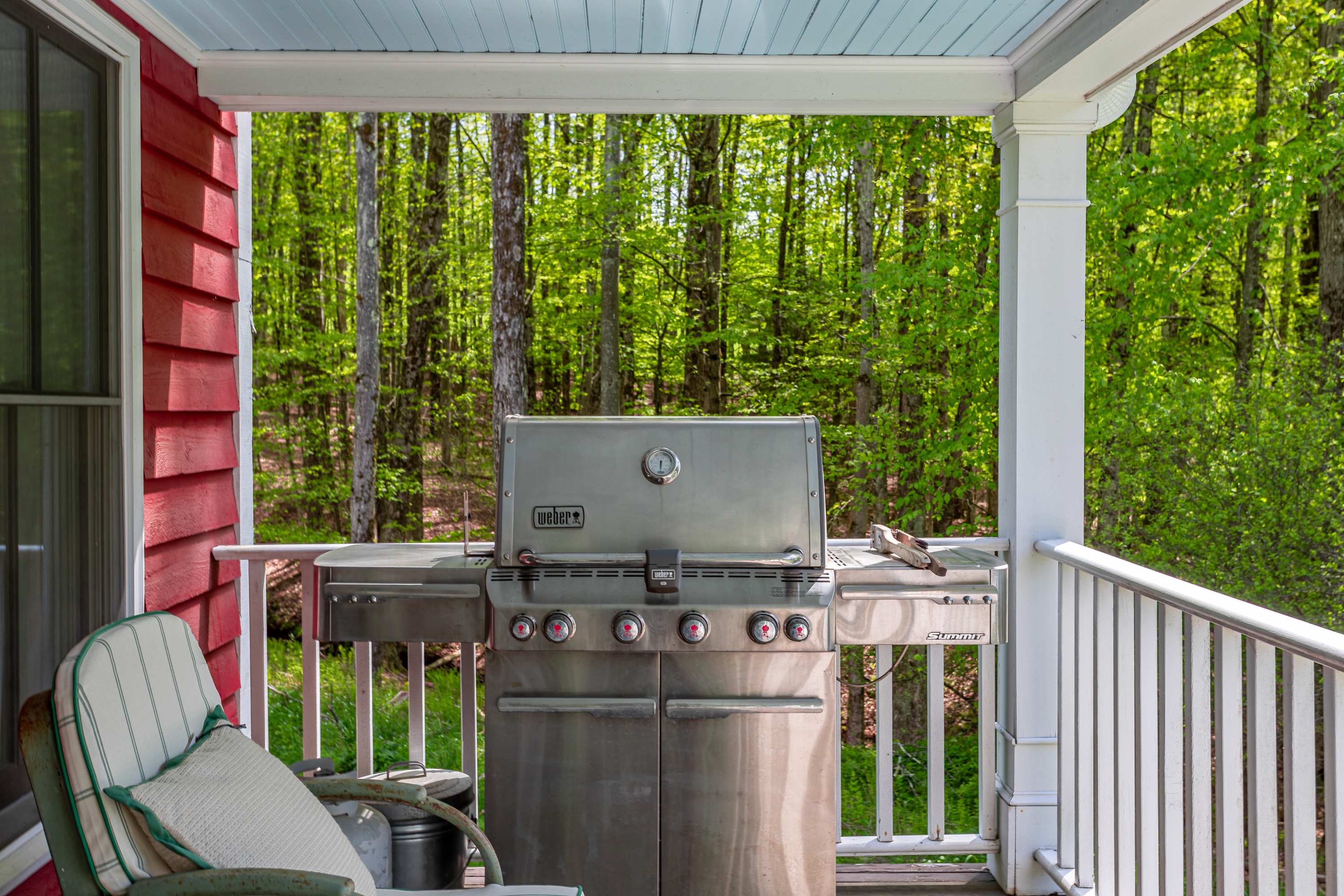 Covered porch area featuring a large stainless steel Weber gas grill, set against a backdrop of vibrant green trees. The porch has dark wood flooring and white railings, perfect for outdoor cooking and entertaining.